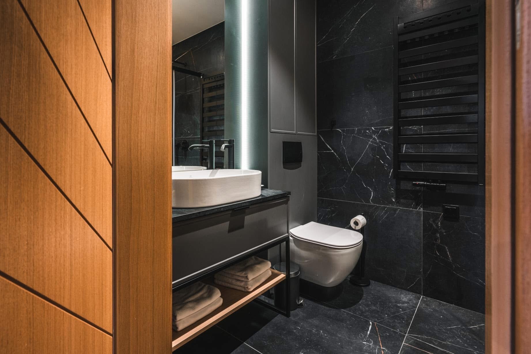 Elegancka, ciemna łazienka z marmurowymi płytkami, owalną umywalką na czarnym blacie i drewnianymi drzwiami z geometrycznym wzorem po lewej stronie.