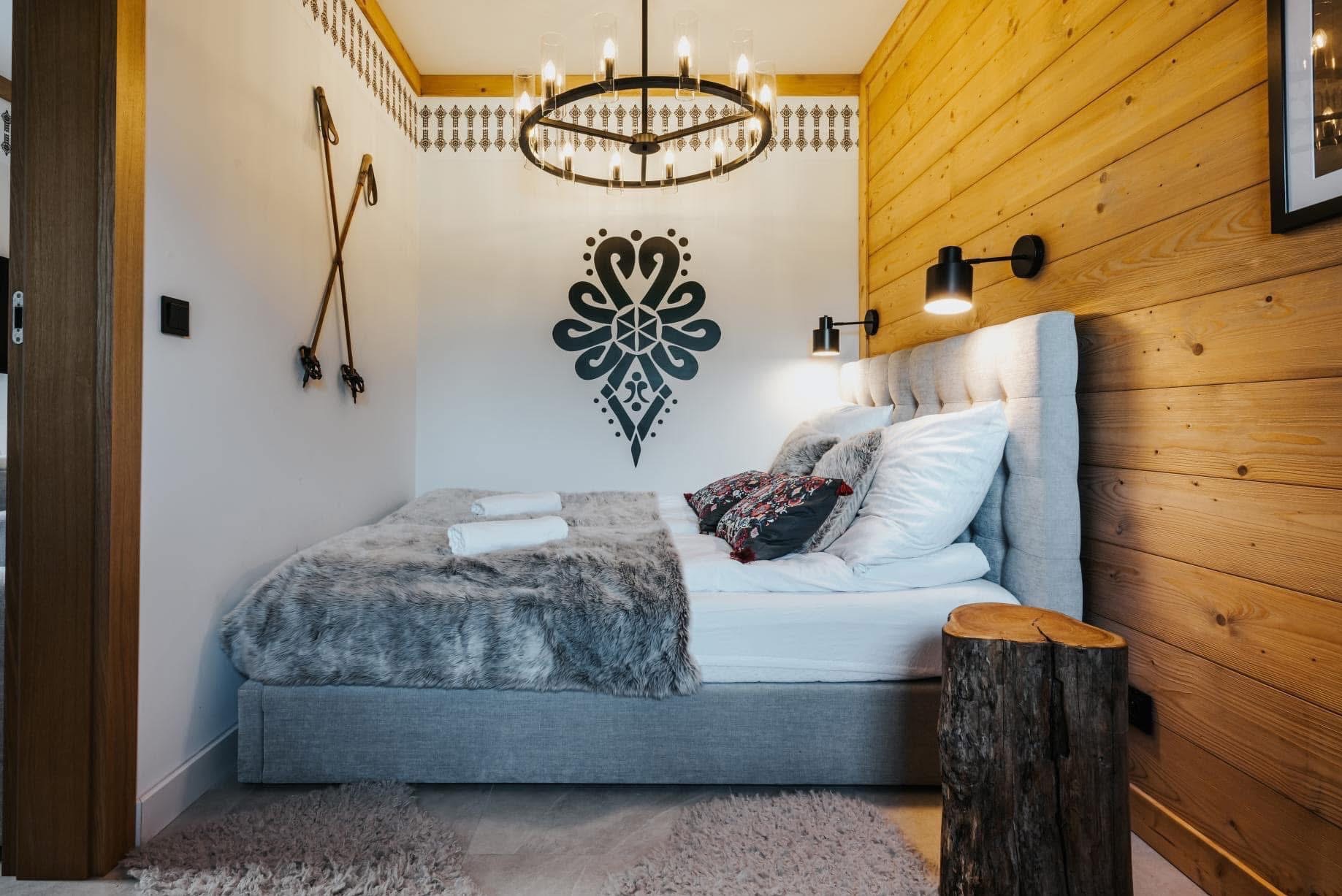 Przytulna sypialnia w góralskim stylu: drewniane panele ścienne, dekoracyjny wzór na ścianie, oryginalne lampy i żyrandol, łóżko z futrzaną narzutą i stolik nocny z pnia.