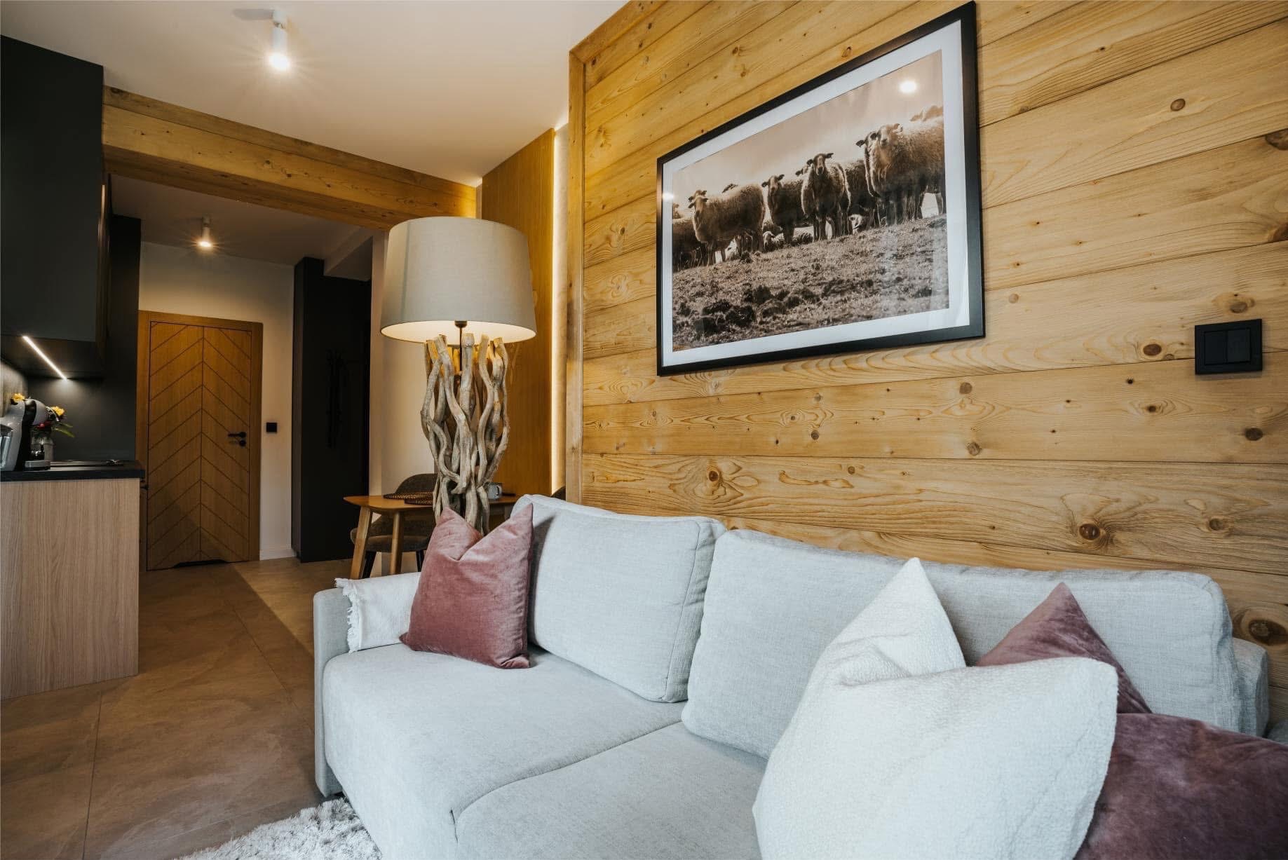 Drewniana ściana z obrazem owiec nad szarą sofą z poduszkami. W tle widoczna kuchnia i drzwi z wzorem w jodełkę. Ciepłe oświetlenie wnętrza.