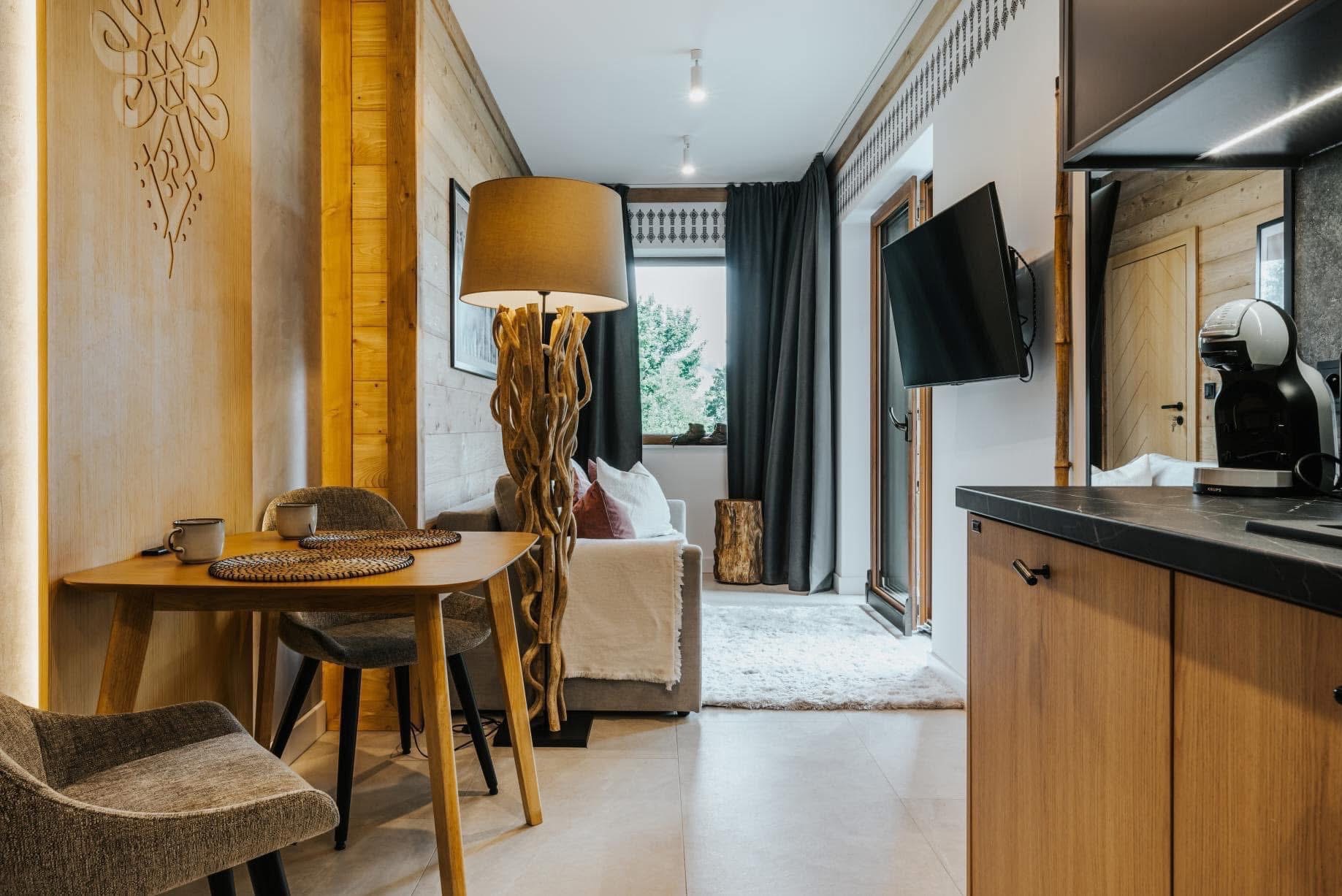 Jasne wnętrze w stylu góralskim: drewniane panele, stół z krzesłami, sofa z poduszkami, unikalna lampa z gałęzi i telewizor na ścianie. Przytulna aranżacja.