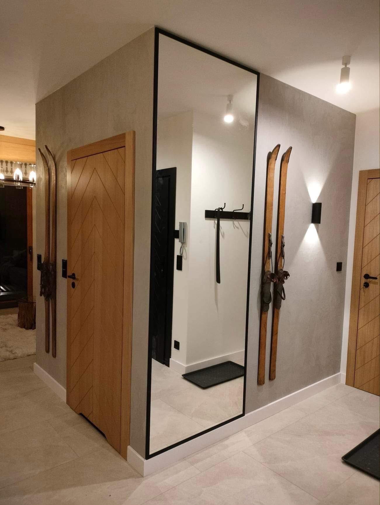 Nowoczesny przedpokój z dużym lustrem w czarnej ramie, drewnianymi drzwiami z wzorem w jodełkę i starymi nartami jako dekoracją ścienną. Minimalistyczne oświetlenie.