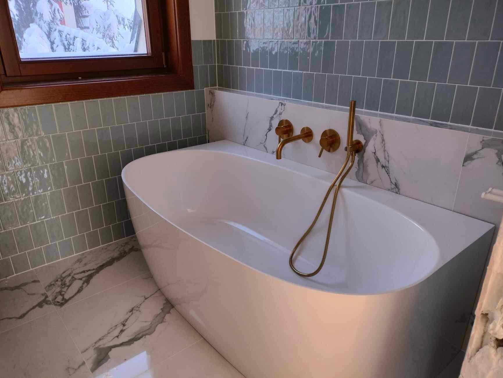 Nowoczesna, wolnostojąca wanna z baterią w kolorze złota na tle ściany wyłożonej szaro-zielonymi płytkami i marmurowym panelem. Podłoga z płytek imitujących marmur.