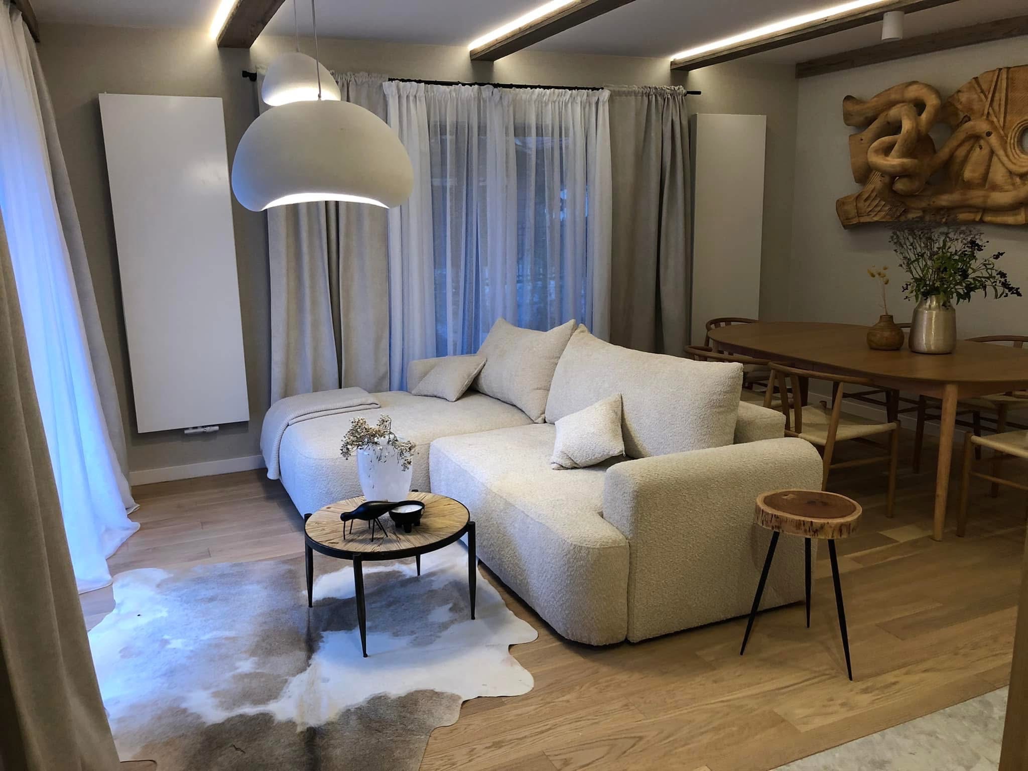 Jasny salon z drewnianą podłogą, beżową sofą i okrągłym stolikiem. Widoczne dekoracje, lampa wisząca i drewniane elementy wykończeniowe sufitu. Przytulne wnętrze.