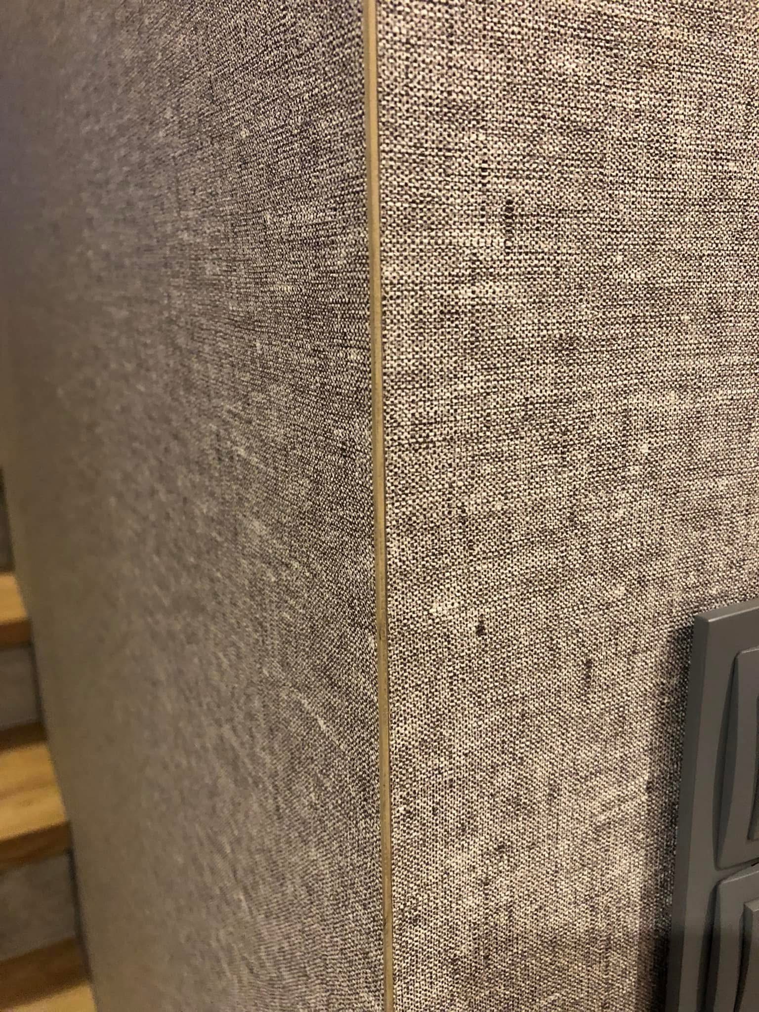 Szczegółowe ujęcie ściany wykończonej tapetą o strukturze lnu w odcieniach szarości, z widocznym narożnikiem i fragmentem włącznika światła.