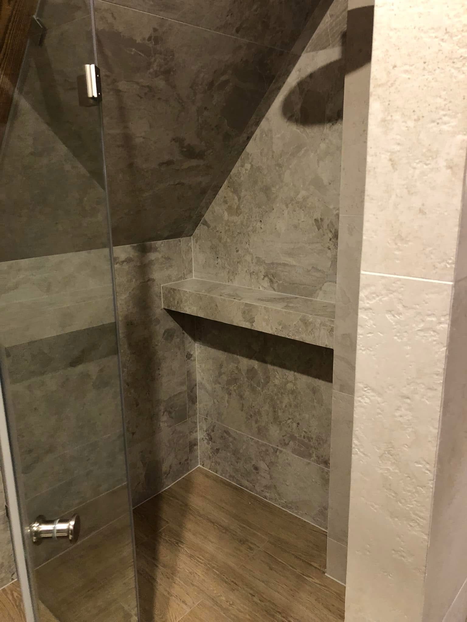 Wykończona kabina prysznicowa w szaro-beżowej tonacji z drewnopodobną podłogą. Widoczna szklana ściana kabiny, półka wewnątrz i płytki imitujące kamień.