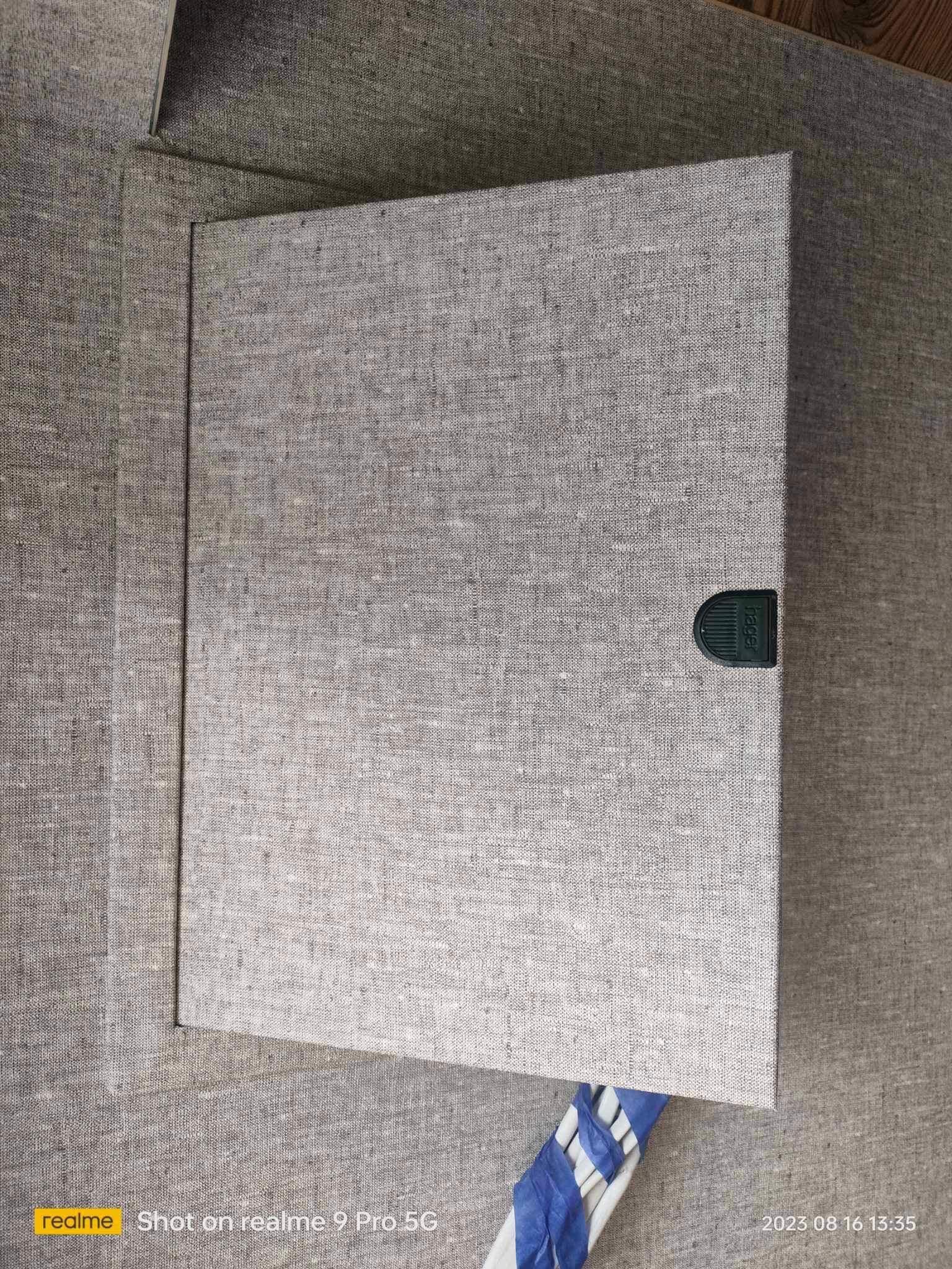 Szary panel inspekcyjny z tkaniny, zamontowany na ścianie, z widocznym zatrzaskiem. Widoczne fragmenty ściany i podłogi.