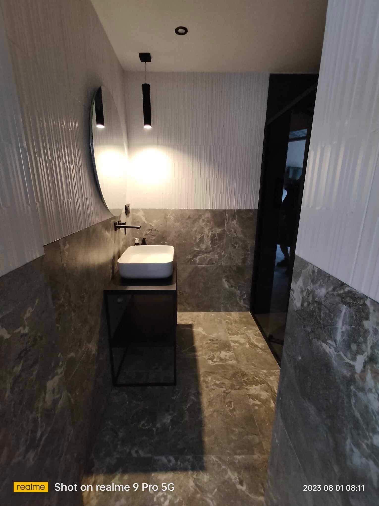 Nowoczesna łazienka po remoncie: biała umywalka nablatowa, czarna armatura, lustro i lampy wiszące. Płytki imitujące marmur na ścianach i podłodze.