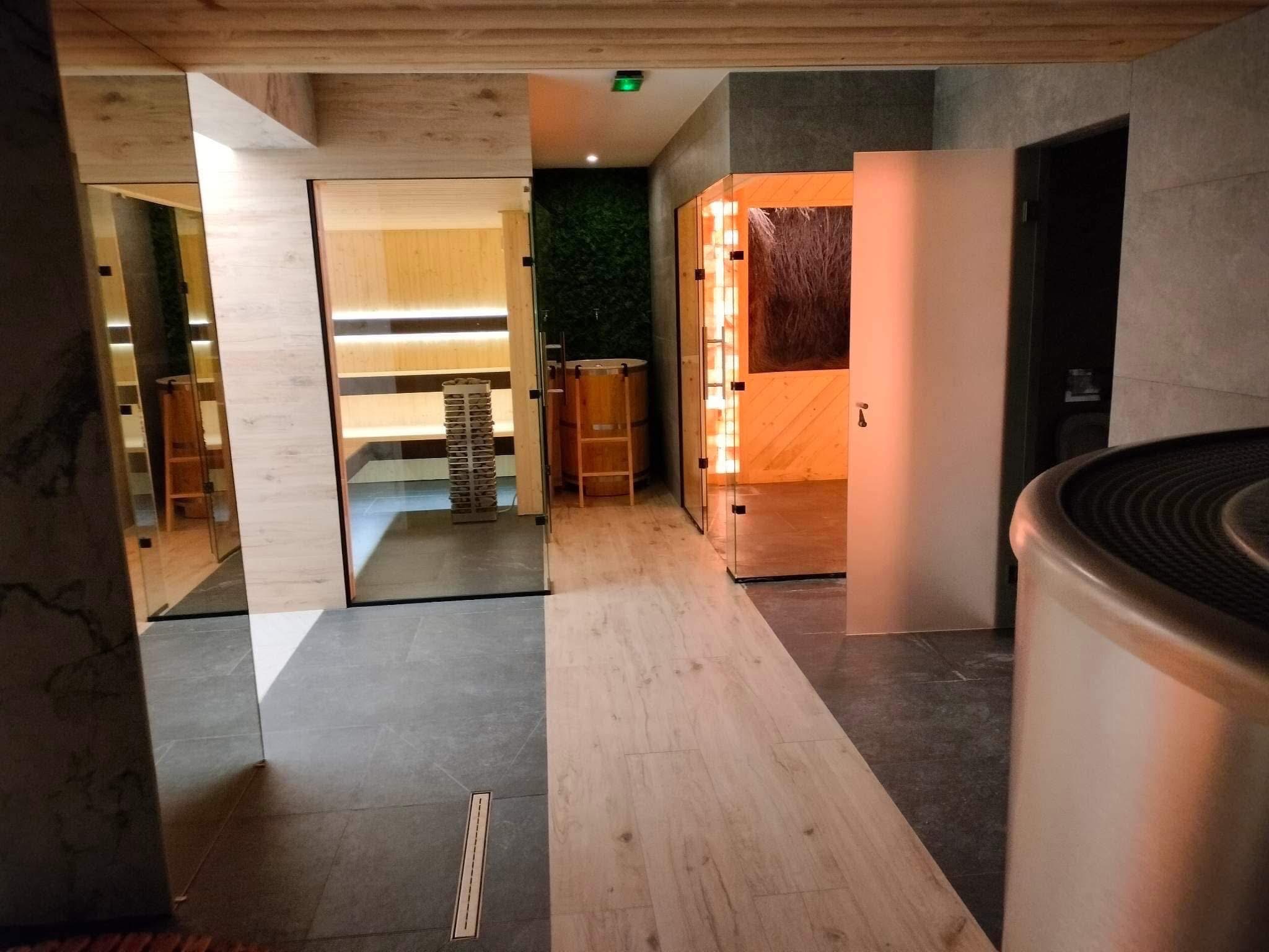 Nowoczesne wnętrze strefy relaksu z saunami: fińska z kamieniami i infrared. Drewniane wykończenie, szklane drzwi, oświetlenie LED, podłoga z płytek w dwóch odcieniach szarości.