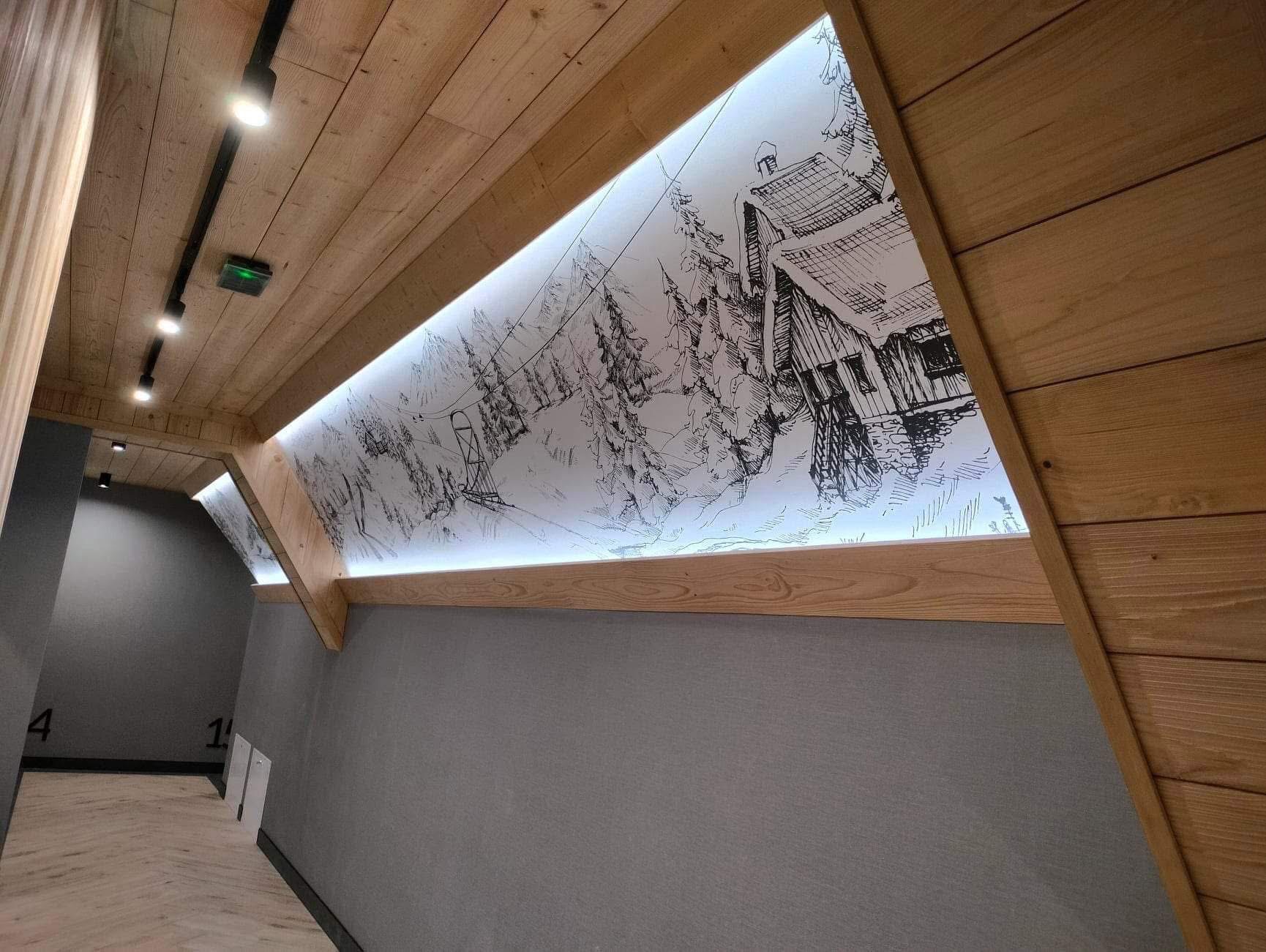 Korytarz z drewnianym sufitem i ścianami, oświetlony rysunkiem górskiego krajobrazu z domkiem i wyciągiem narciarskim. Nowoczesne oświetlenie punktowe.
