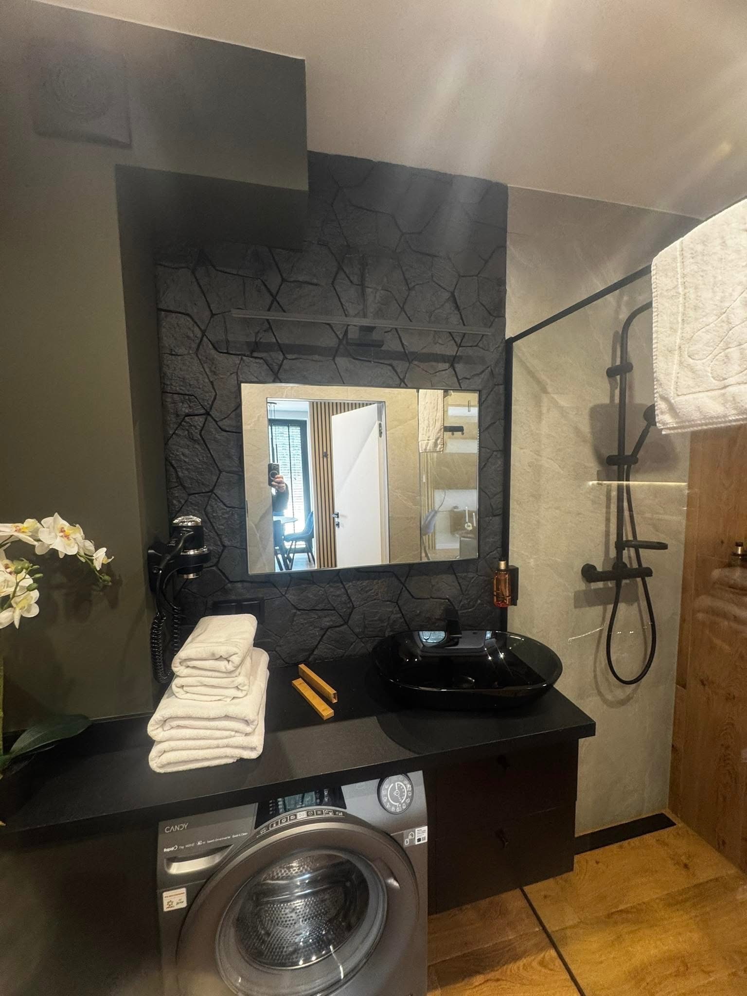 Nowoczesna łazienka z czarną umywalką, kabiną prysznicową i pralką. Ściana za lustrem wykończona kamieniem dekoracyjnym. Widoczne białe ręczniki i kwiaty.