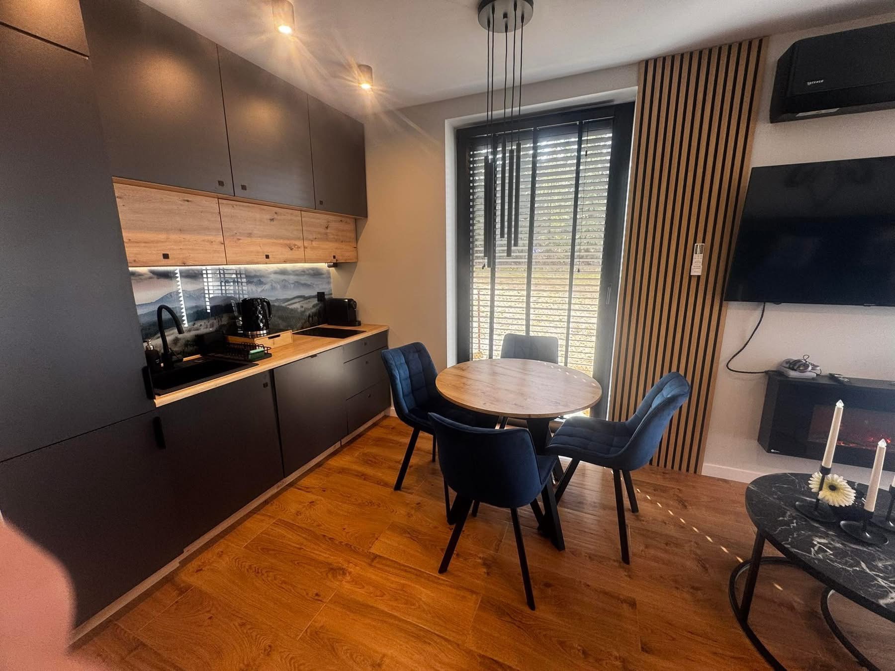 Nowoczesna kuchnia z ciemnymi szafkami, drewnianym blatem i okrągłym stołem z niebieskimi krzesłami. Na ścianie dekoracyjne panele drewniane i telewizor.