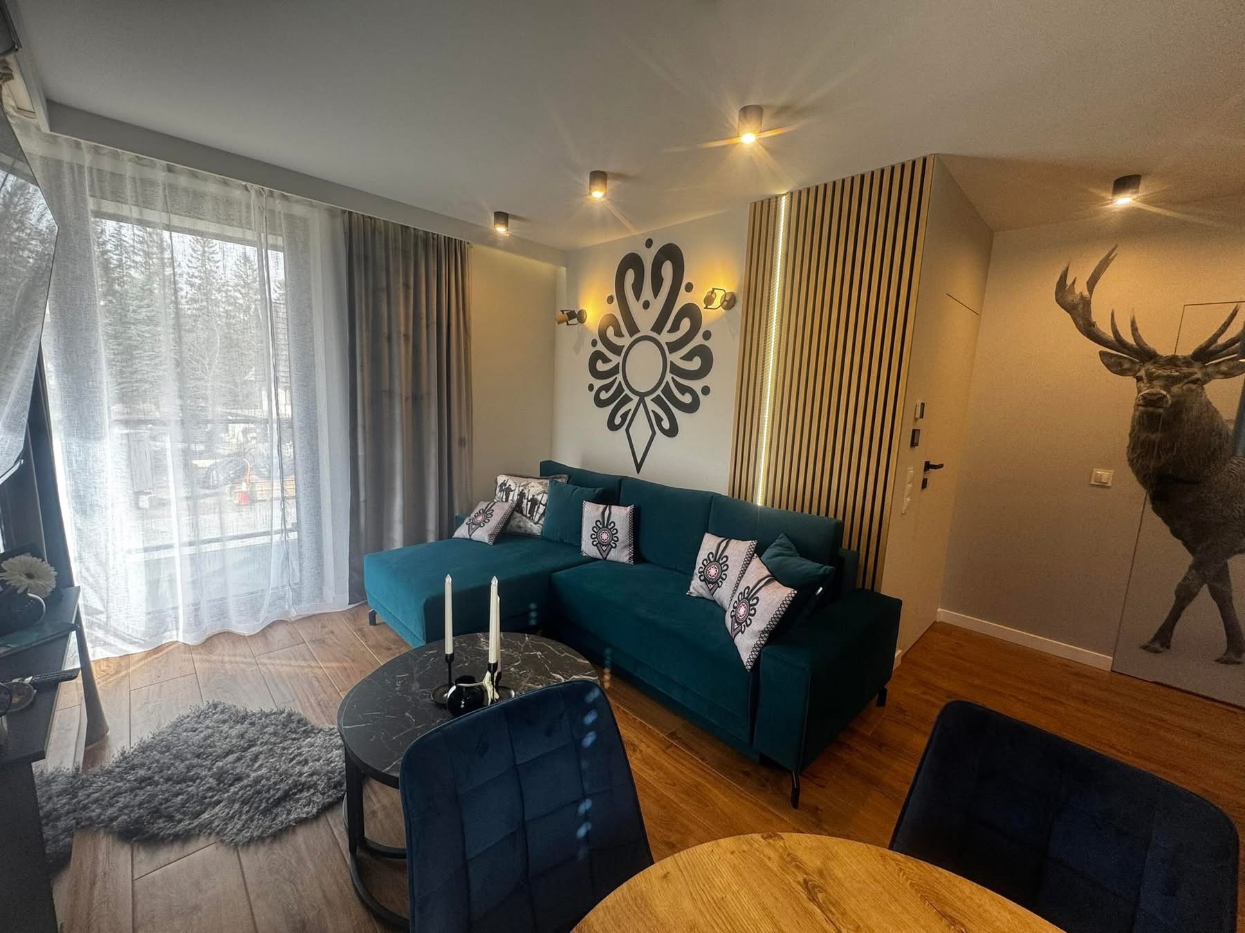 Nowoczesny salon z turkusową sofą narożną, drewnianą boazerią i folkowym motywem na ścianie. Widoczne okno z firanami i drewniana podłoga.