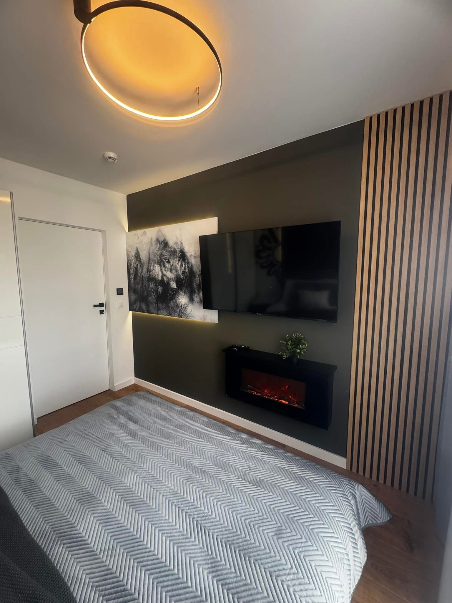 Nowoczesna sypialnia z łóżkiem, panelem ściennym, telewizorem i elektrycznym kominkiem. Ciemna ściana z obrazem wilka podświetlonym ledami. Okrągła lampa sufitowa.