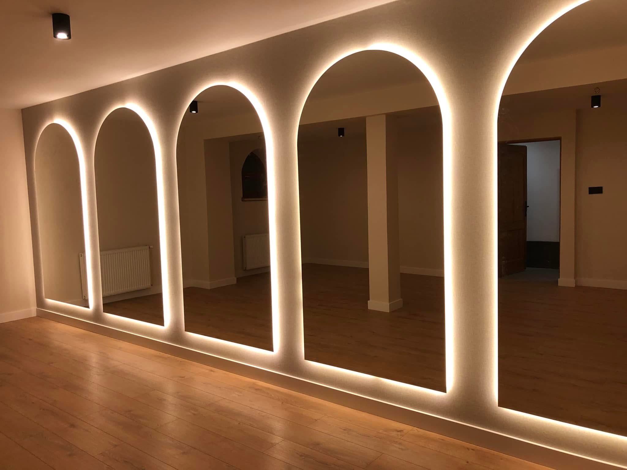 Nowoczesne wnętrze z podświetlanymi wnękami w kształcie łuków, odbijającymi się w lustrach. Parkiet z jasnego drewna, minimalistyczne oświetlenie punktowe.
