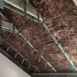 Serhii Toniuk - Izolacja poddasza z wełny mineralnej, mocowana do drewnianej konstrukcji za pomocą metalowych uchwytów i sznurka. Widok z dołu na ukośną powierzchnię.