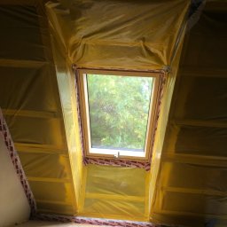 Serhii Toniuk - Okno dachowe zabezpieczone folią malarską w trakcie prac wykończeniowych poddasza. Widoczne drewniane elementy konstrukcyjne i surowa podłoga.