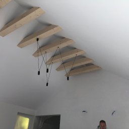 Serhii Toniuk - Biały sufit z drewnianymi belkami i minimalistycznymi lampami wiszącymi. Widoczne ślady po szpachlowaniu na ścianach. Ujęcie z dołu, fragment osoby w prawym dolnym rogu.