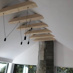 Serhii Toniuk - Białe wnętrze poddasza z drewnianymi belkami i czarnymi lampami wiszącymi. Fragment surowego komina z cegieł. Okna z widokiem na zieleń.