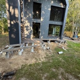 Serhii Toniuk - Montaż drewnianego tarasu przy nowoczesnym domu z szarą elewacją. Widoczne betonowe słupy i drewniana konstrukcja nośna. W tle drzewa i kontener.