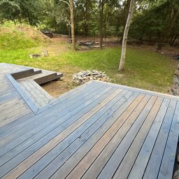 Serhii Toniuk - Nowoczesny taras z desek kompozytowych w odcieniach szarości, z niskimi schodami i widokiem na zielony ogród z drzewami. W tle stos drewna i hamak.