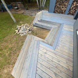 Serhii Toniuk - Nowoczesny taras drewniany z szarych desek, schody z drewna, widok z góry na tle zieleni i drzew. Ułożone drewno opałowe przy ścianie budynku.