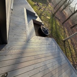 Serhii Toniuk - Szara deska tarasowa z widocznymi schodkami, otoczona zielenią i drzewami w słoneczny dzień. Widok z góry, z fragmentem krzesła i buta w kadrze.