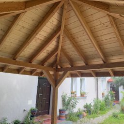 Serhii Toniuk - Drewniana konstrukcja altany z widocznym dachem z desek i elementami nośnymi. W tle budynek mieszkalny z oknami i drzwiami, roślinność w donicach.