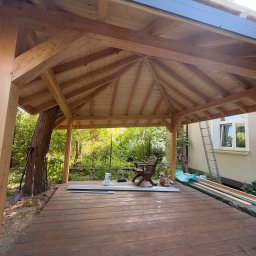 Serhii Toniuk - Drewniana altana w ogrodzie, widok od wewnątrz. Konstrukcja z belek, desek i zadaszenia. W tle zieleń drzew i krzewów, drabina oparta o ścianę budynku.