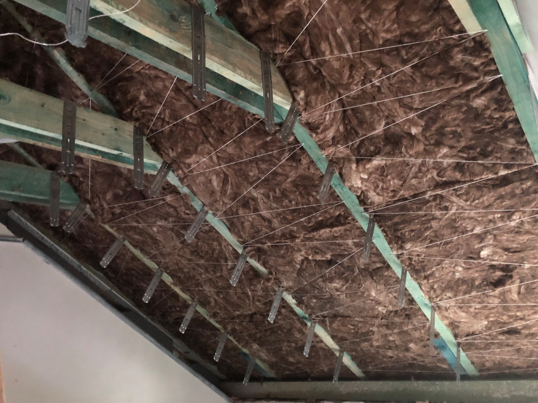 Izolacja poddasza z wełny mineralnej, mocowana do drewnianej konstrukcji za pomocą metalowych uchwytów i sznurka. Widok z dołu na ukośną powierzchnię.