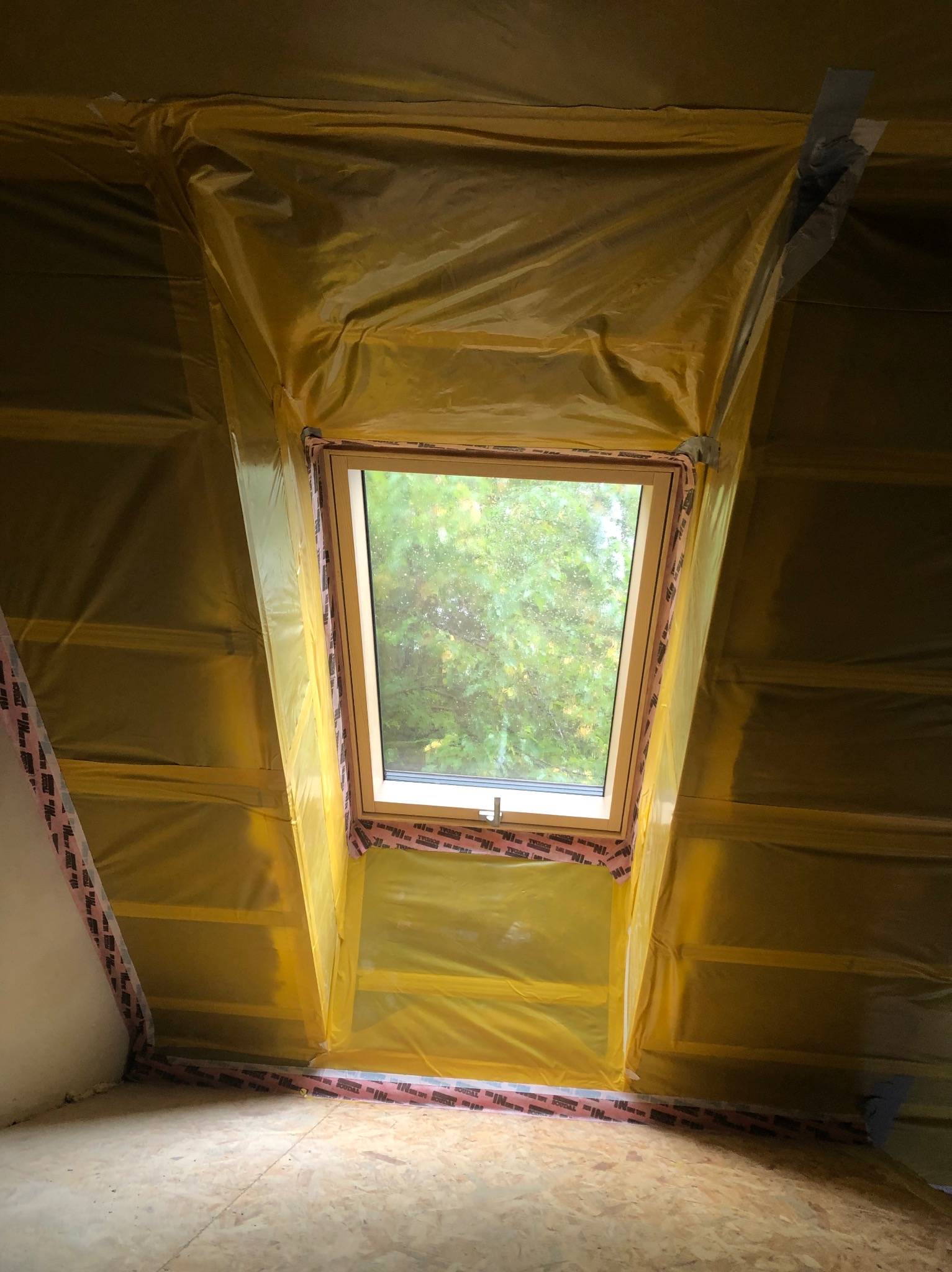 Okno dachowe zabezpieczone folią malarską w trakcie prac wykończeniowych poddasza. Widoczne drewniane elementy konstrukcyjne i surowa podłoga.