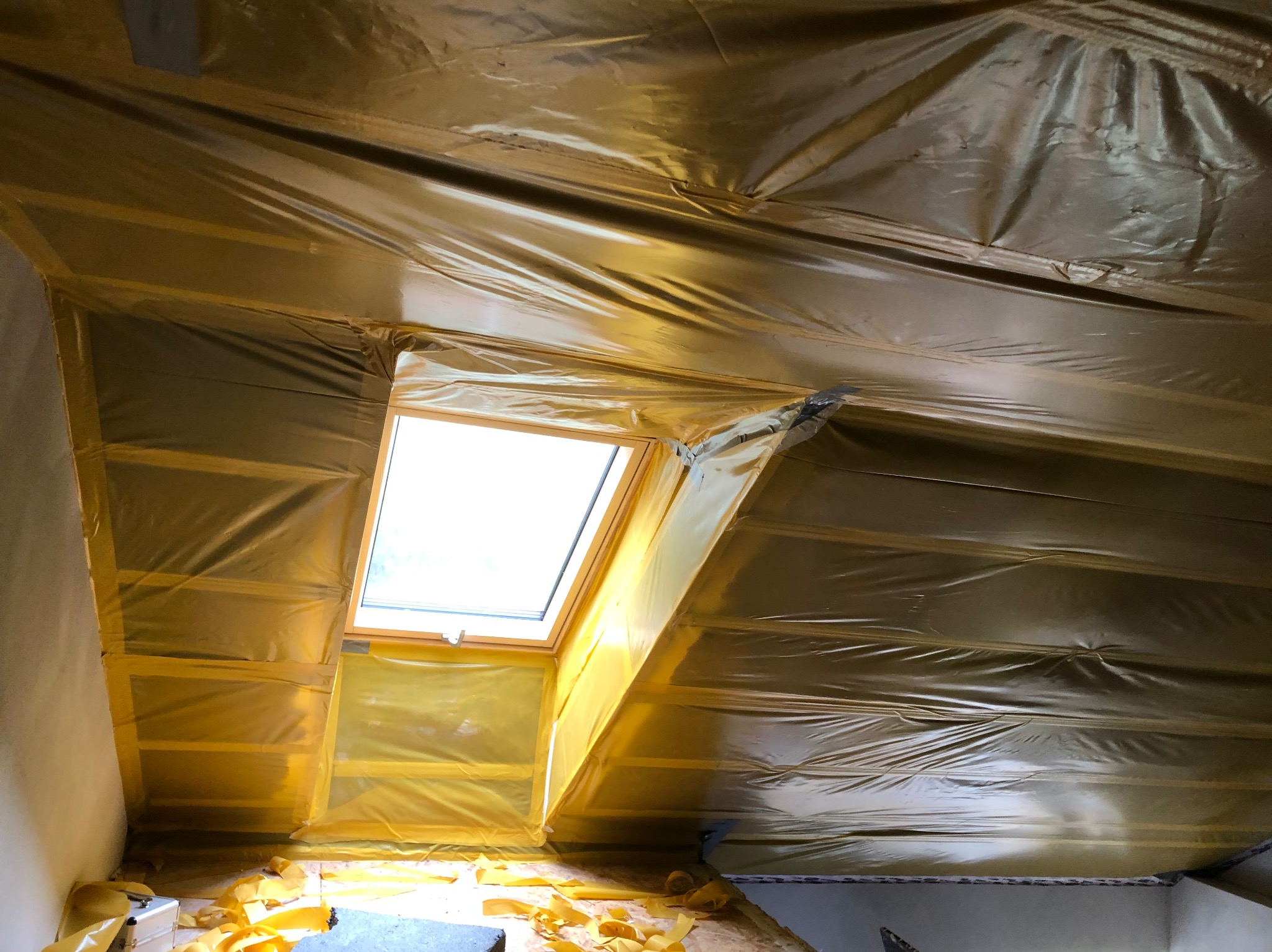 Skośny sufit poddasza z oknem dachowym, zabezpieczony folią w kolorze żółtym, przygotowany do dalszych prac wykończeniowych. Widoczne drewniane elementy konstrukcyjne.