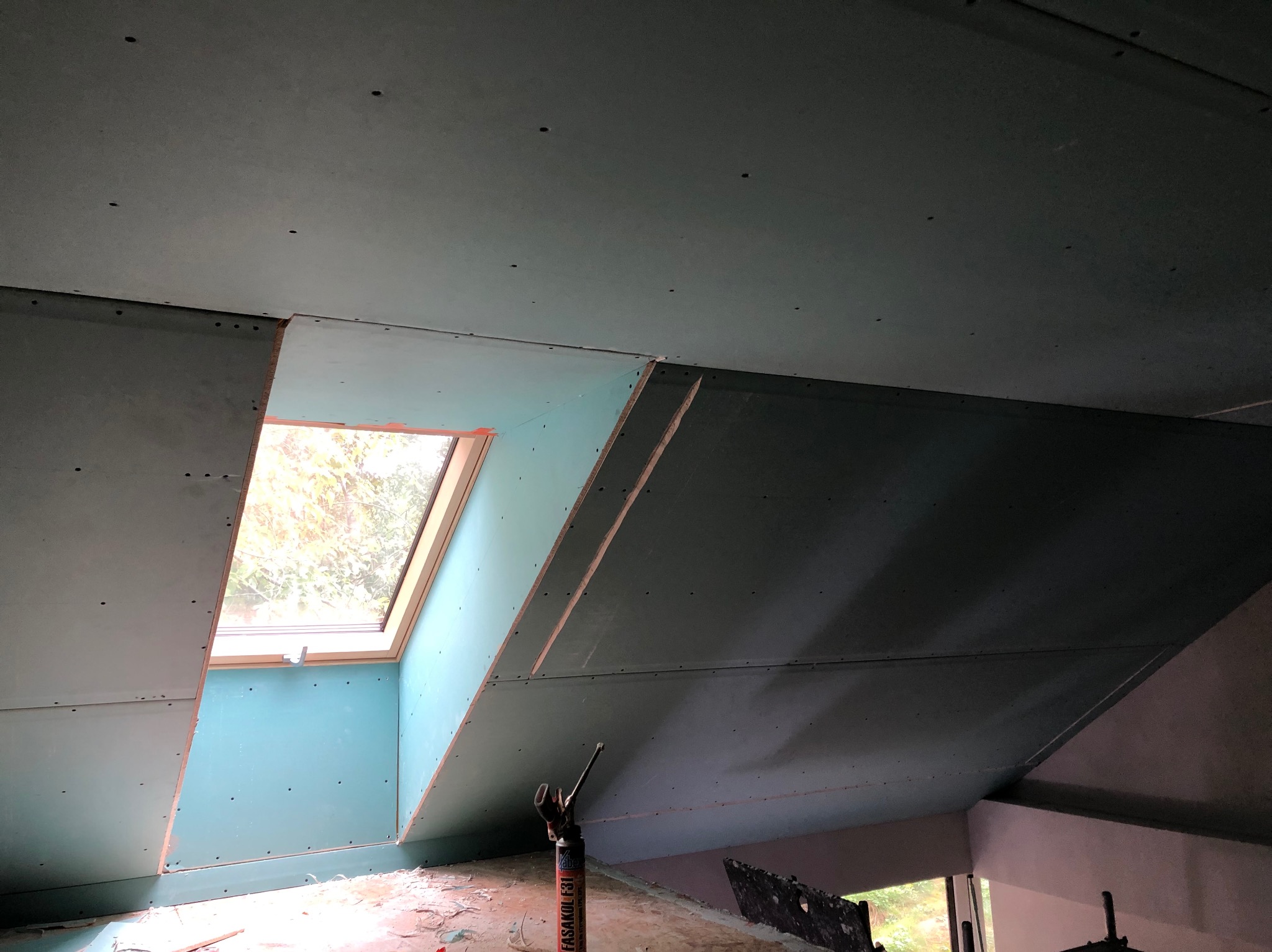 Montaż płyty G-K na poddaszu z oknem dachowym. Widoczne narzędzia i fragment ściany. Płyty w kolorze zielonym, sufit skośny.