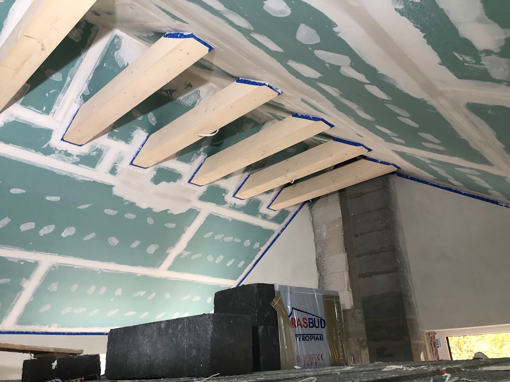 Skośny sufit z płyt gipsowo-kartonowych w trakcie montażu, widoczne drewniane belki i taśma malarska. Częściowo zaszpachlowane łączenia płyt.