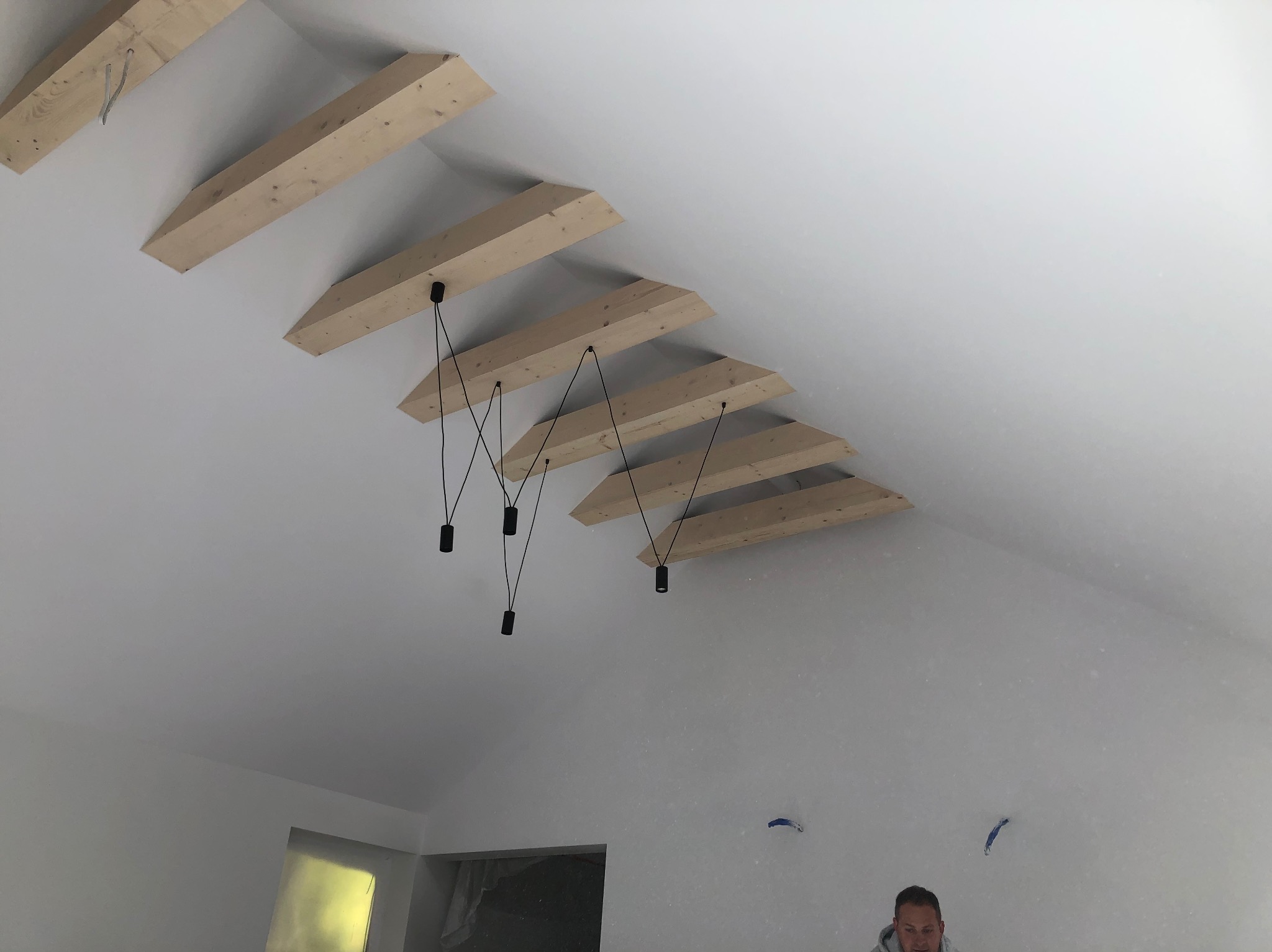 Biały sufit z drewnianymi belkami i minimalistycznymi lampami wiszącymi. Widoczne ślady po szpachlowaniu na ścianach. Ujęcie z dołu, fragment osoby w prawym dolnym rogu.