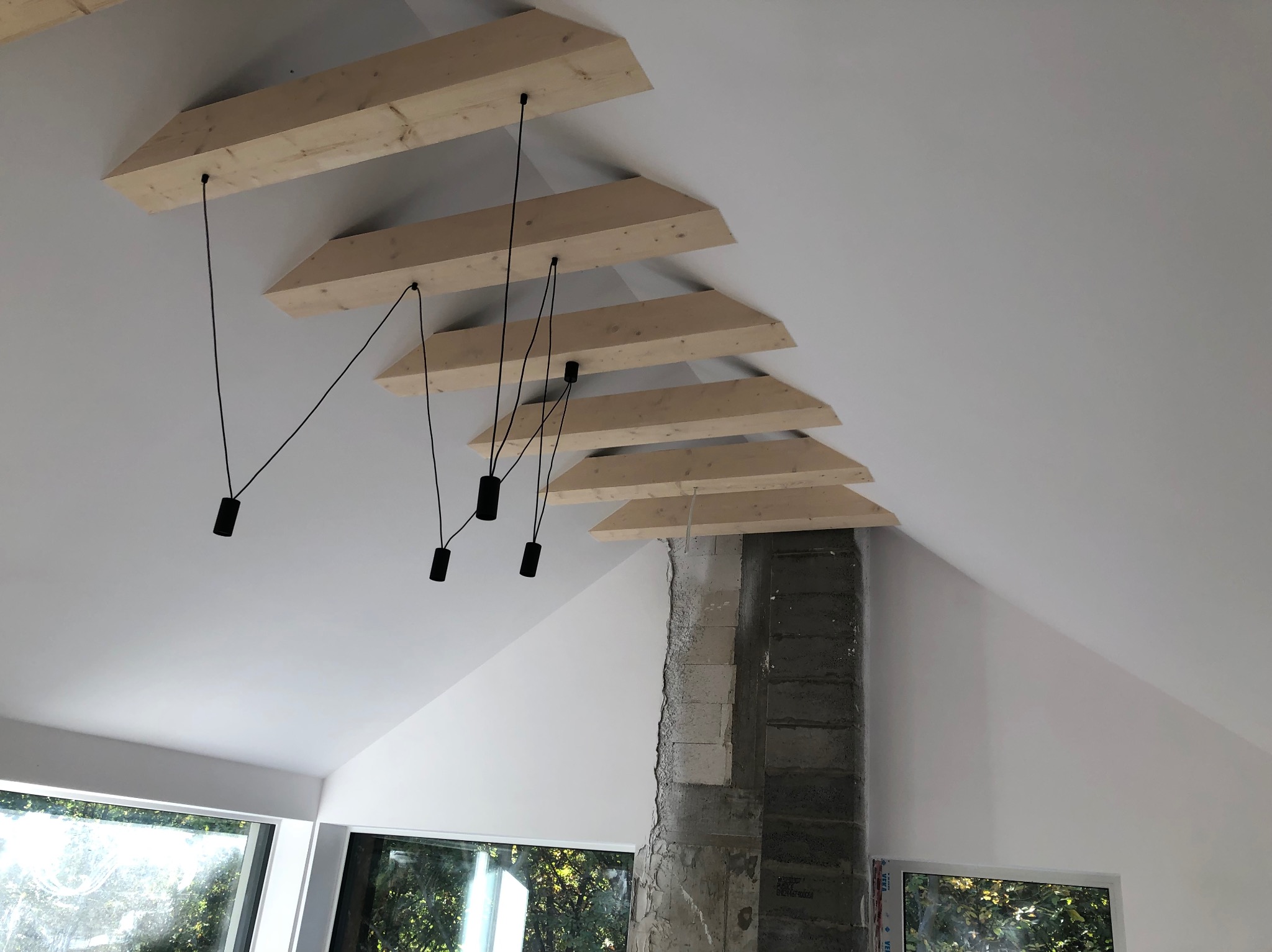 Białe wnętrze poddasza z drewnianymi belkami i czarnymi lampami wiszącymi. Fragment surowego komina z cegieł. Okna z widokiem na zieleń.