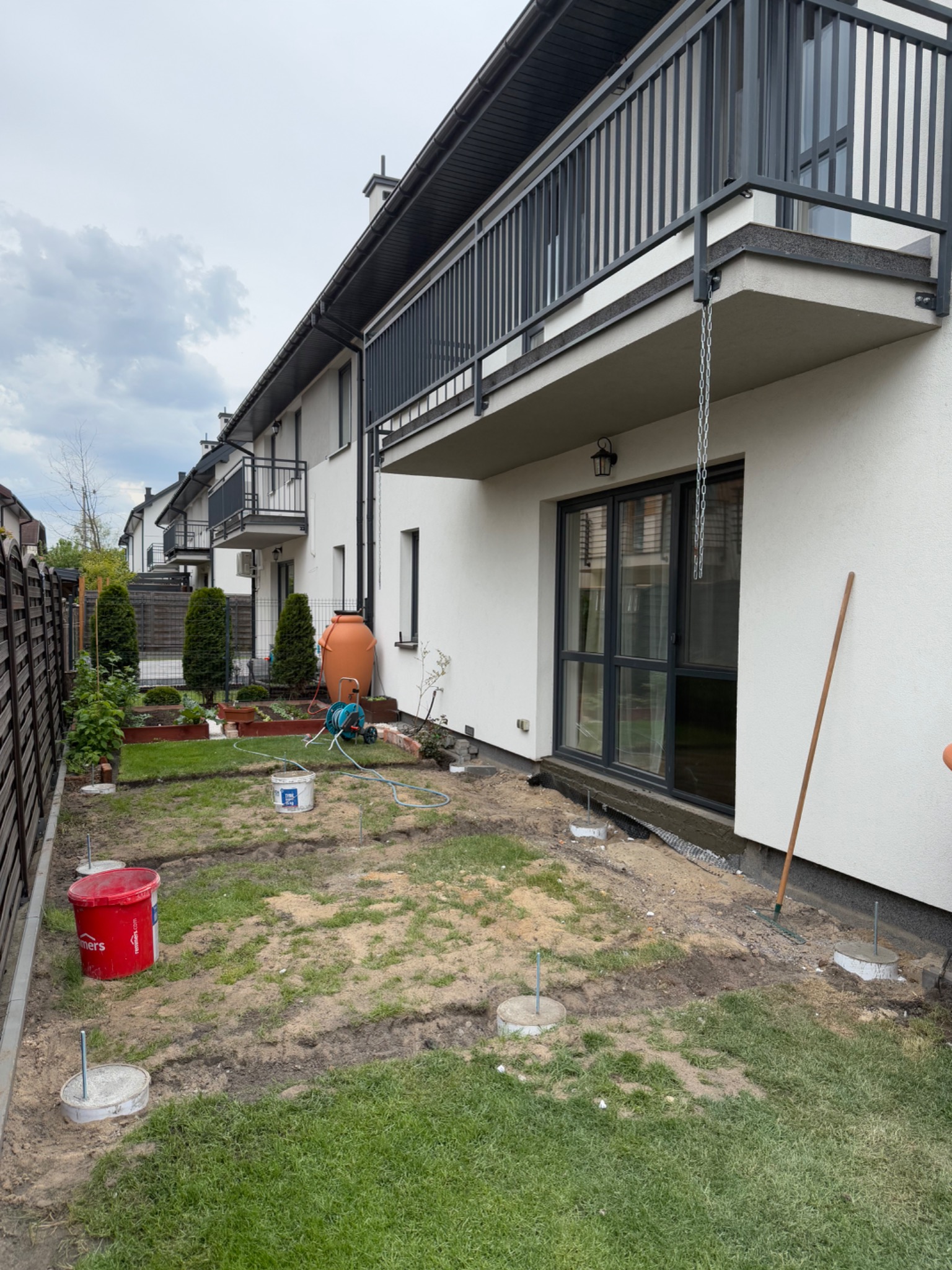 Przygotowanie podłoża pod taras drewniany: widoczne betonowe podstawy, wytyczone obrysy, narzędzia i materiały budowlane na trawniku przy nowoczesnym domu.