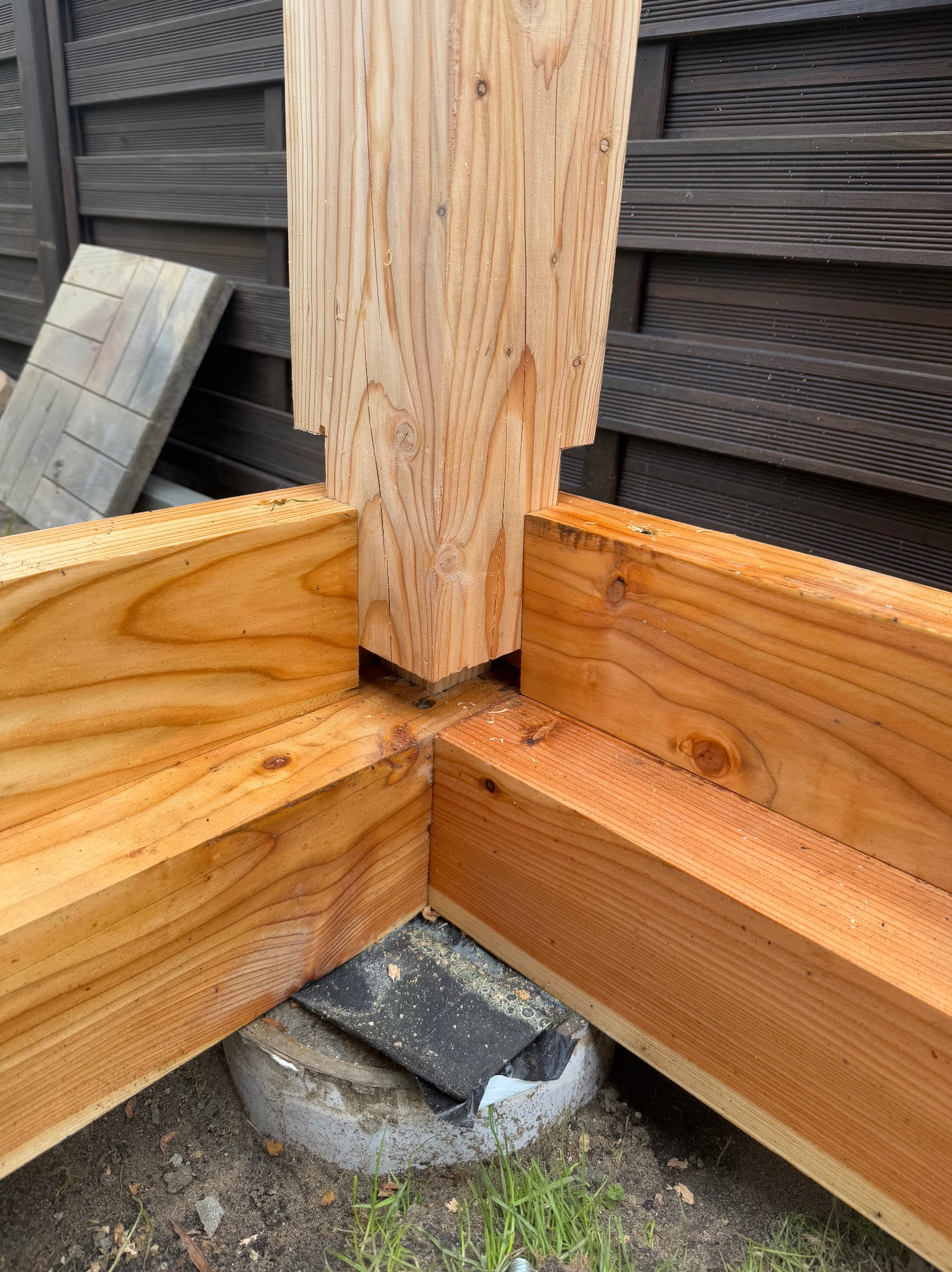 Drewniana konstrukcja tarasu w trakcie budowy, widoczne połączenia belek i fundament. W tle brązowe deski ogrodzenia.