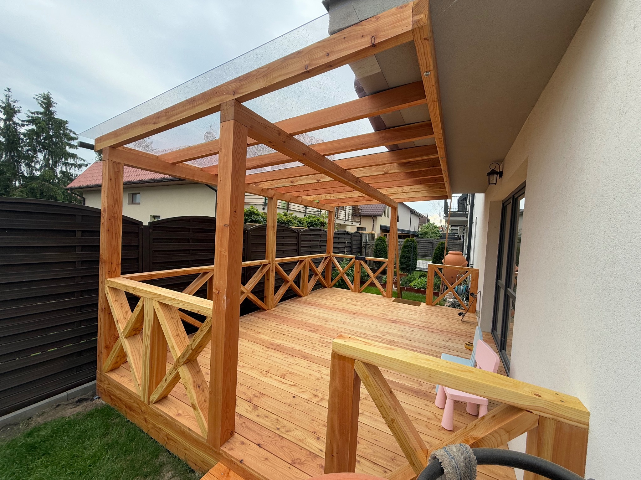 Zadaszony taras drewniany z balustradą krzyżową, przylegający do budynku. Widoczne dziecięce krzesełka i fragment ogrodu. Całość w ciepłej tonacji drewna.