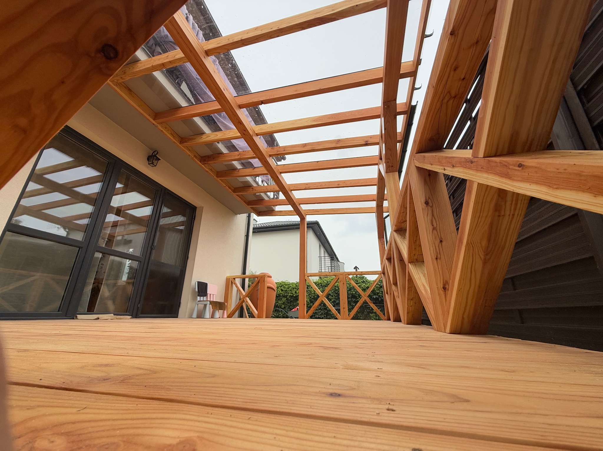 Nowoczesny taras drewniany z zadaszeniem, widok z dołu, ukazujący strukturę drewna i detale konstrukcyjne. Jasne drewno kontrastuje z ciemnymi ramami okien.