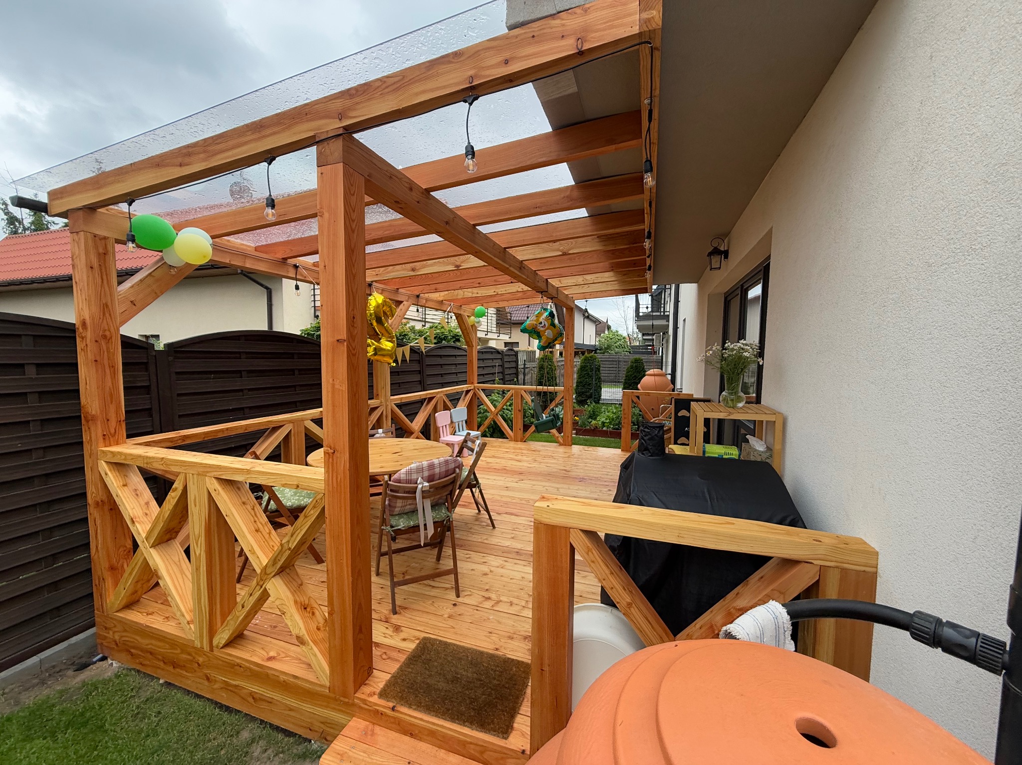 Drewniany taras z zadaszeniem i balustradą, udekorowany balonami. Widoczny stół z krzesłami i elementy wyposażenia. Całość w jasnych, naturalnych odcieniach drewna.