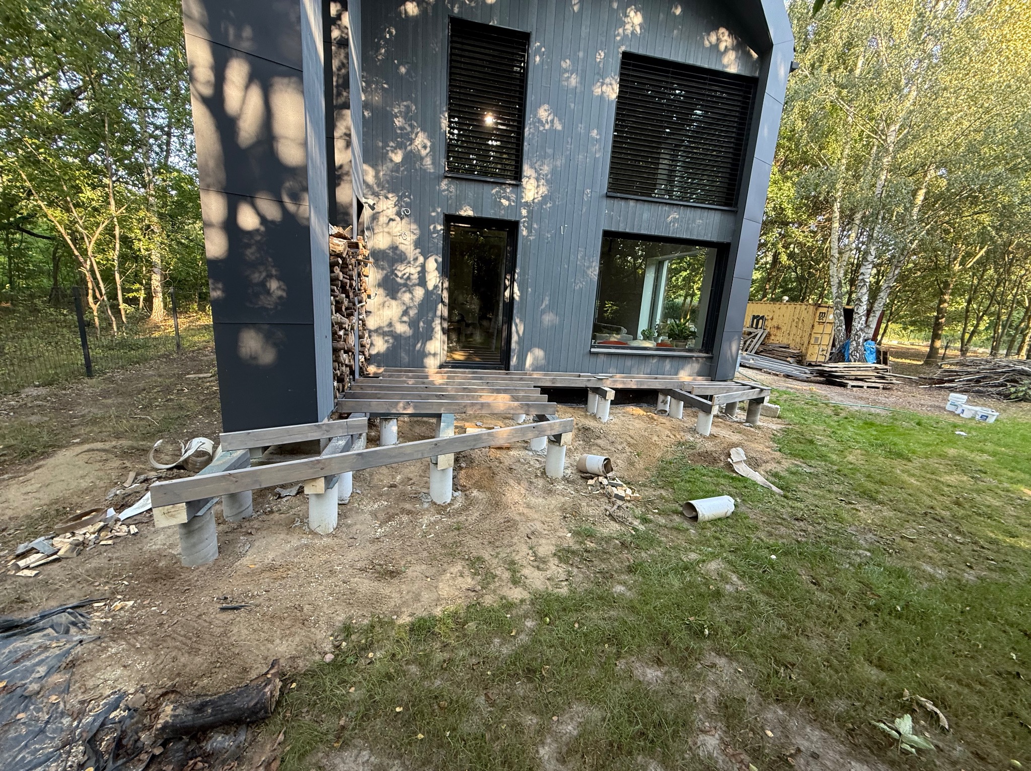 Montaż drewnianego tarasu przy nowoczesnym domu z szarą elewacją. Widoczne betonowe słupy i drewniana konstrukcja nośna. W tle drzewa i kontener.