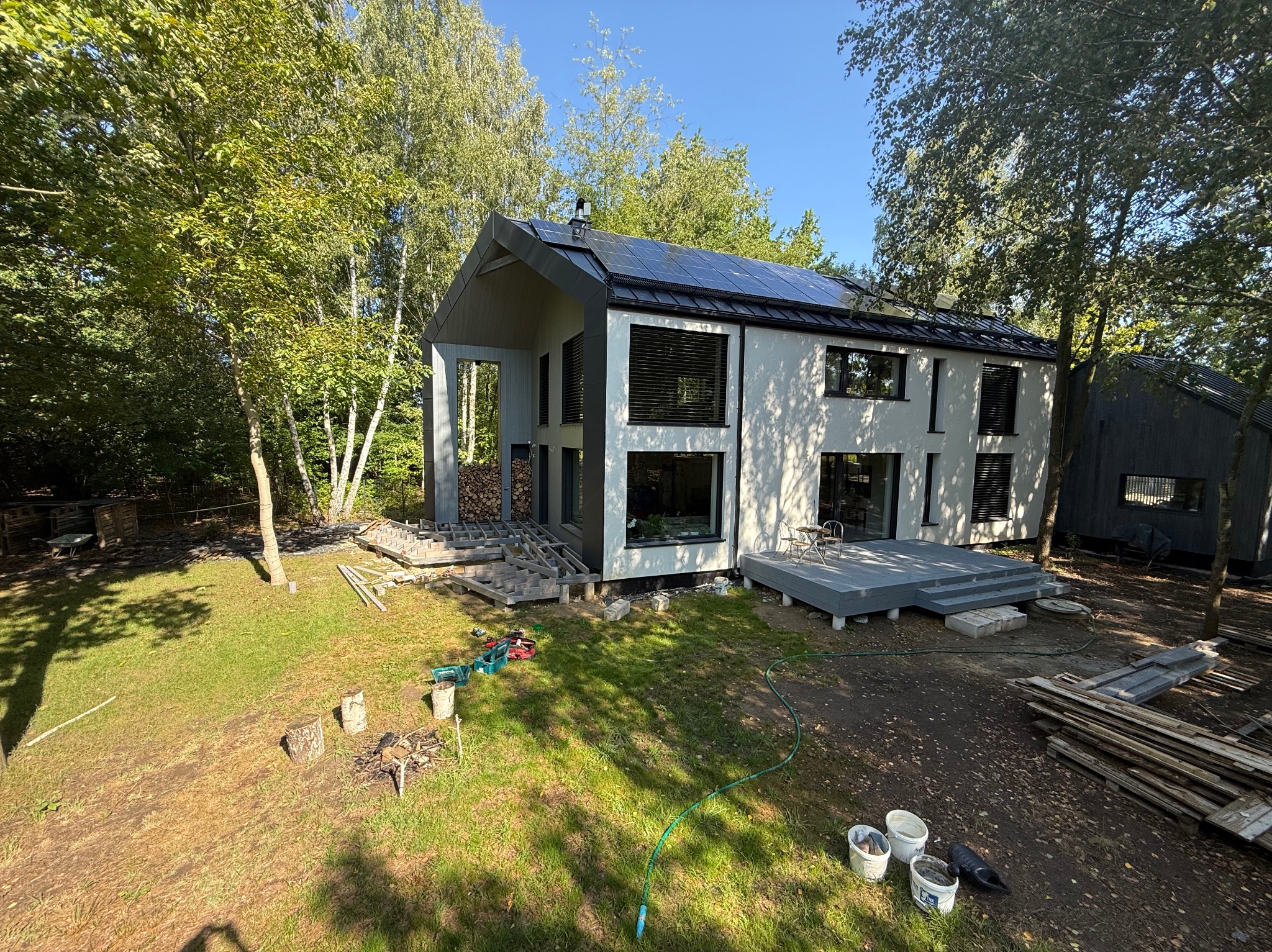 Nowoczesny dom z panelami słonecznymi i szarym tarasem w trakcie budowy, otoczony zielenią. Widoczne narzędzia i materiały budowlane.