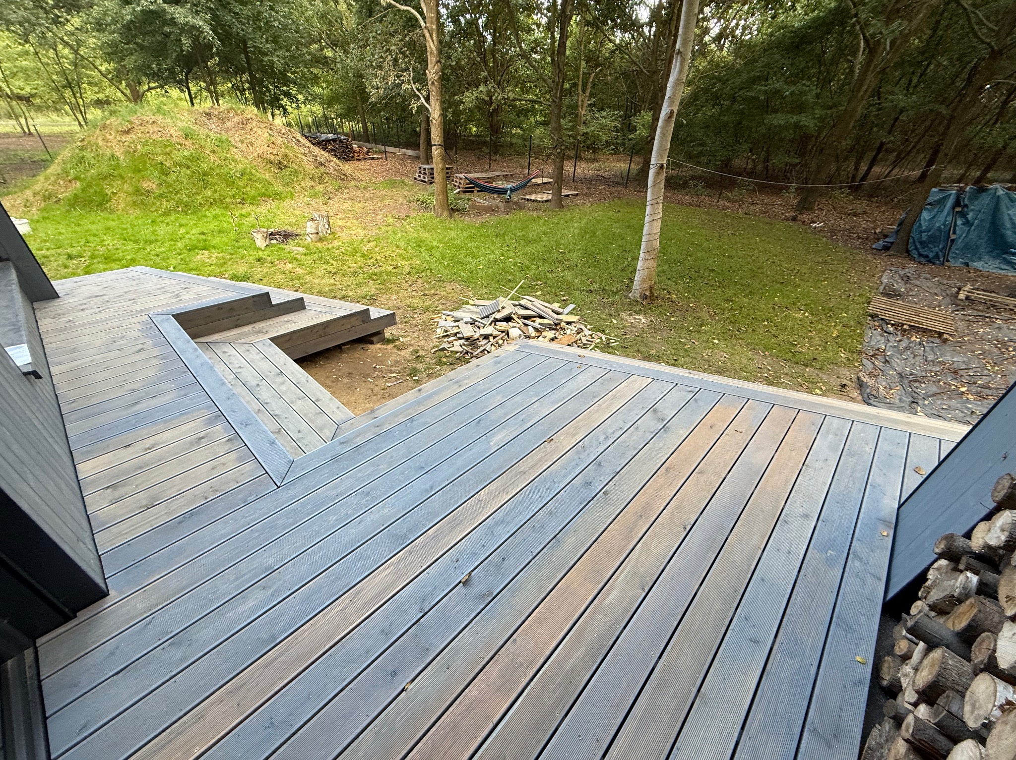 Nowoczesny taras z desek kompozytowych w odcieniach szarości, z niskimi schodami i widokiem na zielony ogród z drzewami. W tle stos drewna i hamak.