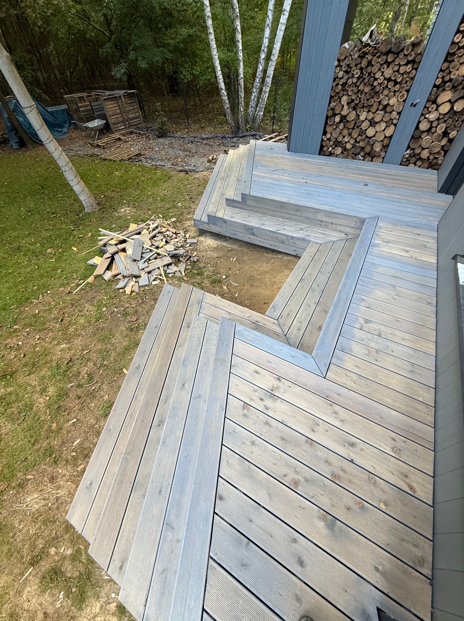 Nowoczesny taras drewniany z szarych desek, schody z drewna, widok z góry na tle zieleni i drzew. Ułożone drewno opałowe przy ścianie budynku.