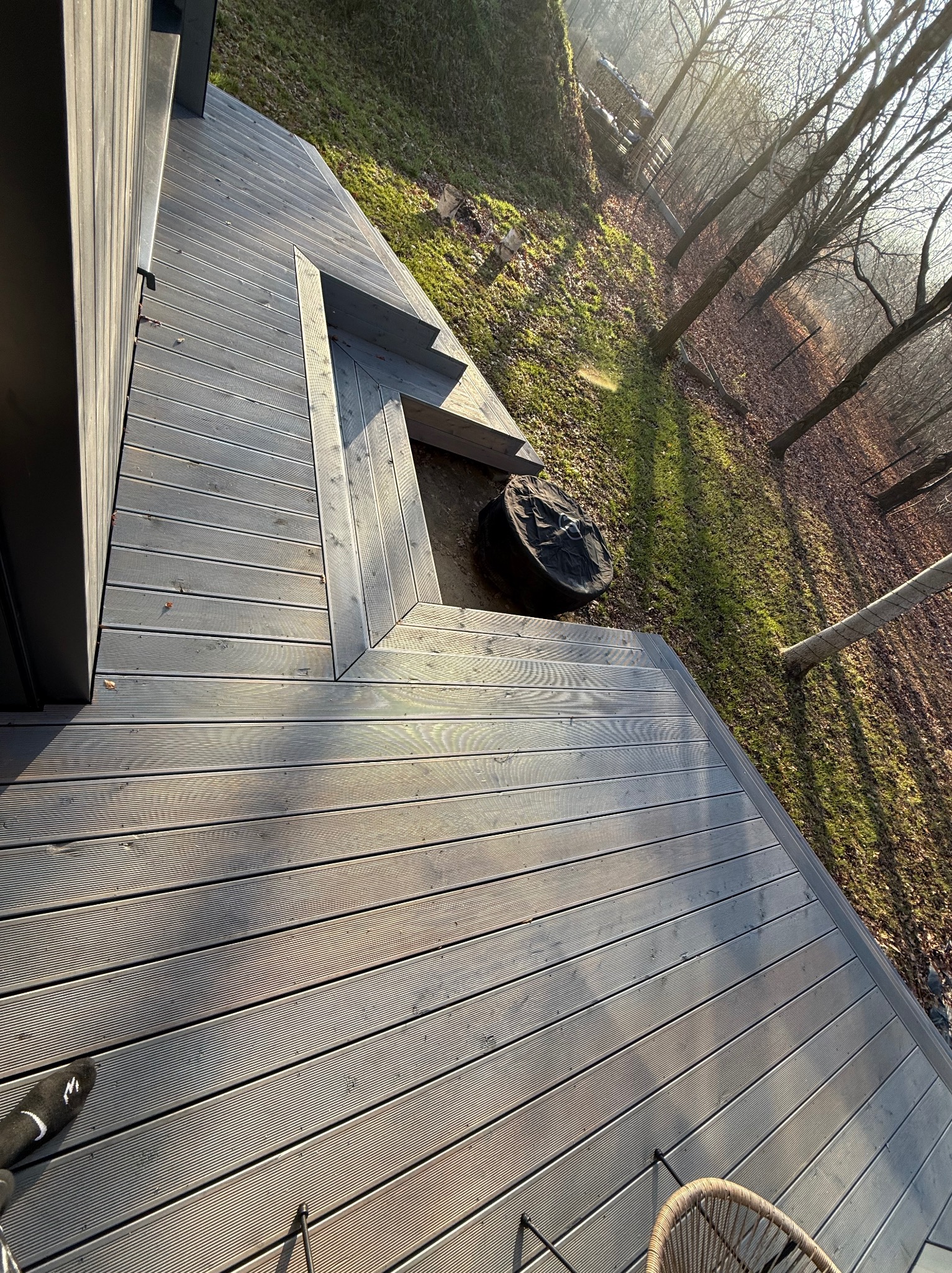 Szara deska tarasowa z widocznymi schodkami, otoczona zielenią i drzewami w słoneczny dzień. Widok z góry, z fragmentem krzesła i buta w kadrze.