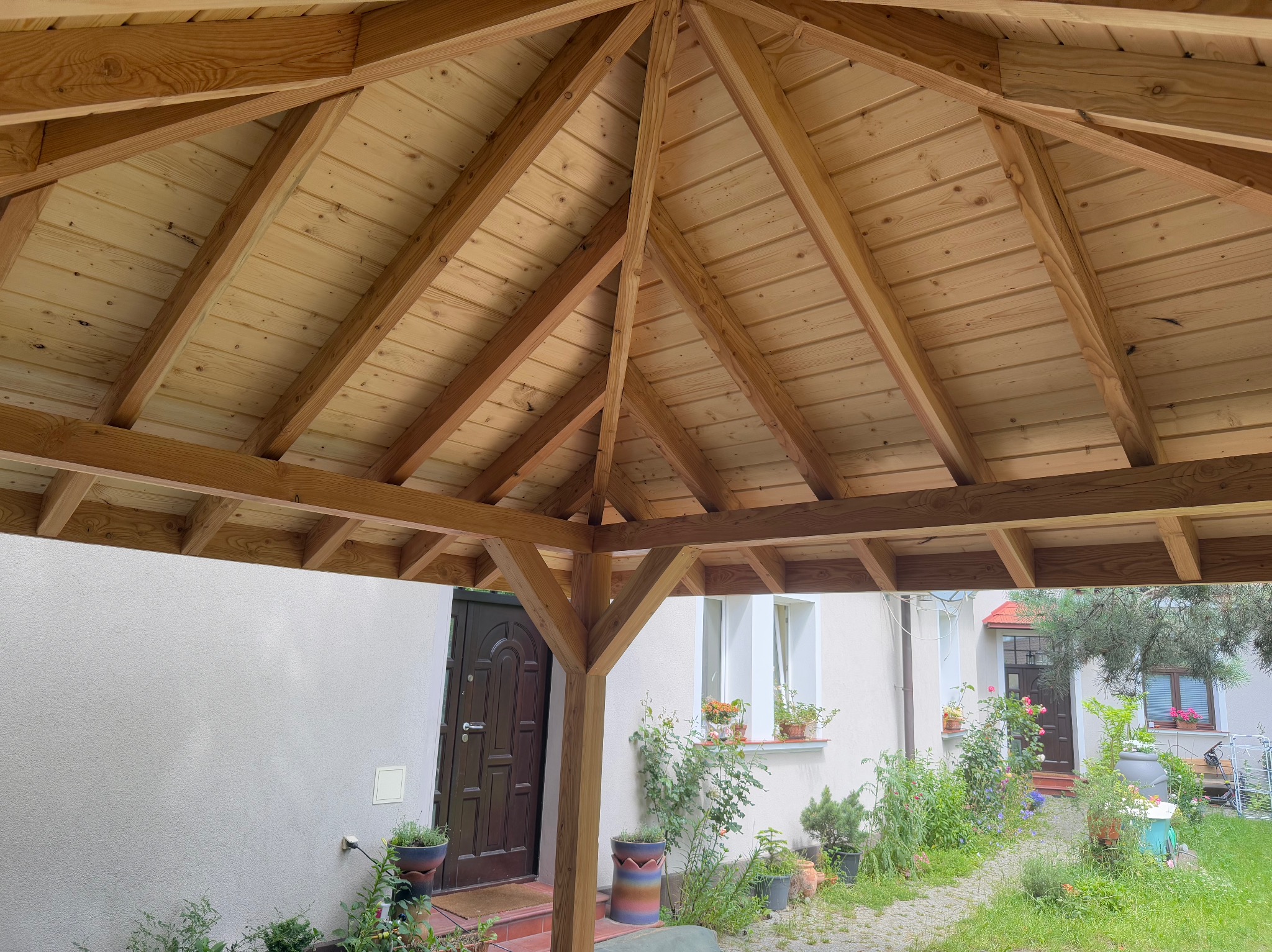Drewniana konstrukcja altany z widocznym dachem z desek i elementami nośnymi. W tle budynek mieszkalny z oknami i drzwiami, roślinność w donicach.