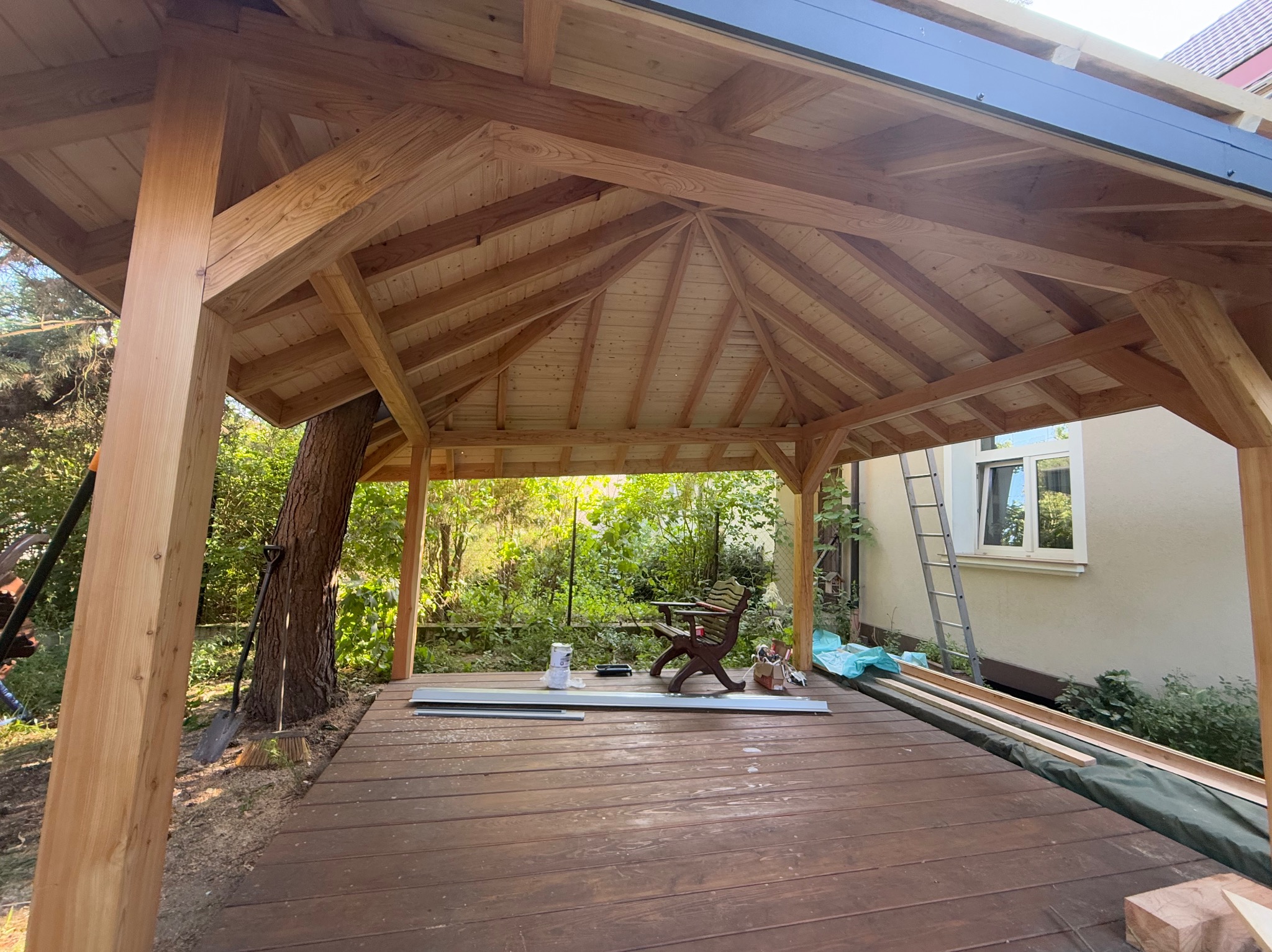 Drewniana altana w ogrodzie, widok od wewnątrz. Konstrukcja z belek, desek i zadaszenia. W tle zieleń drzew i krzewów, drabina oparta o ścianę budynku.