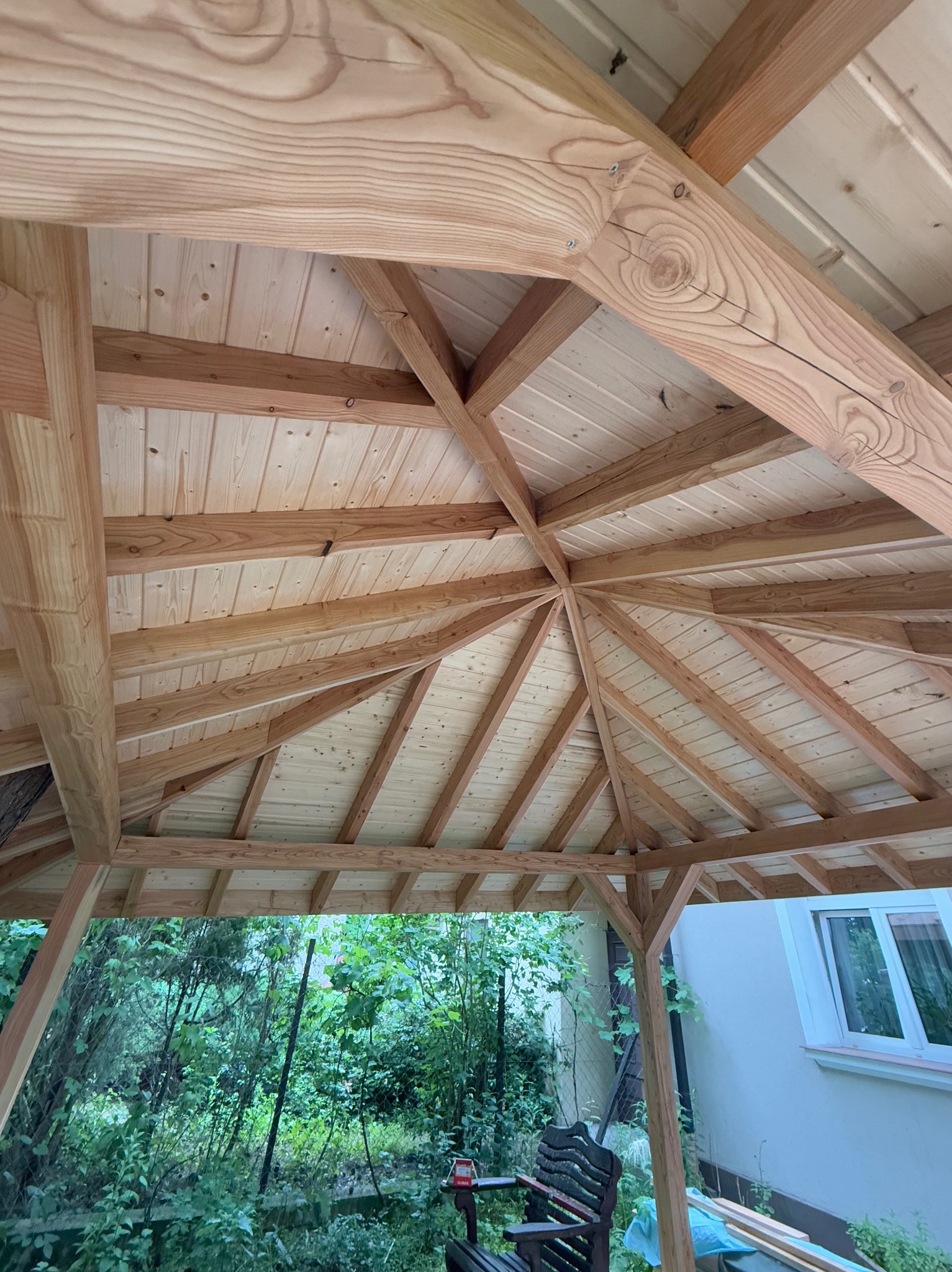 Drewniana konstrukcja altany z widocznym dachem i belkami, z zewnątrz widok na zieleń ogrodu i fragment budynku z oknem. Jasne drewno, symetryczny układ belek.