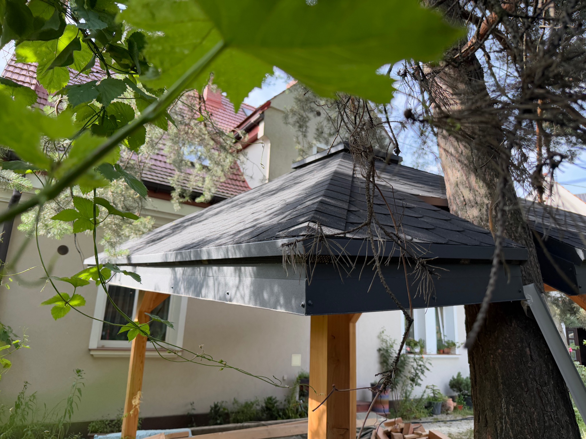 Szara altana z drewnianymi słupami w ogrodzie, widoczna spod liści winorośli. W tle dom z czerwoną dachówką i drzewo iglaste.