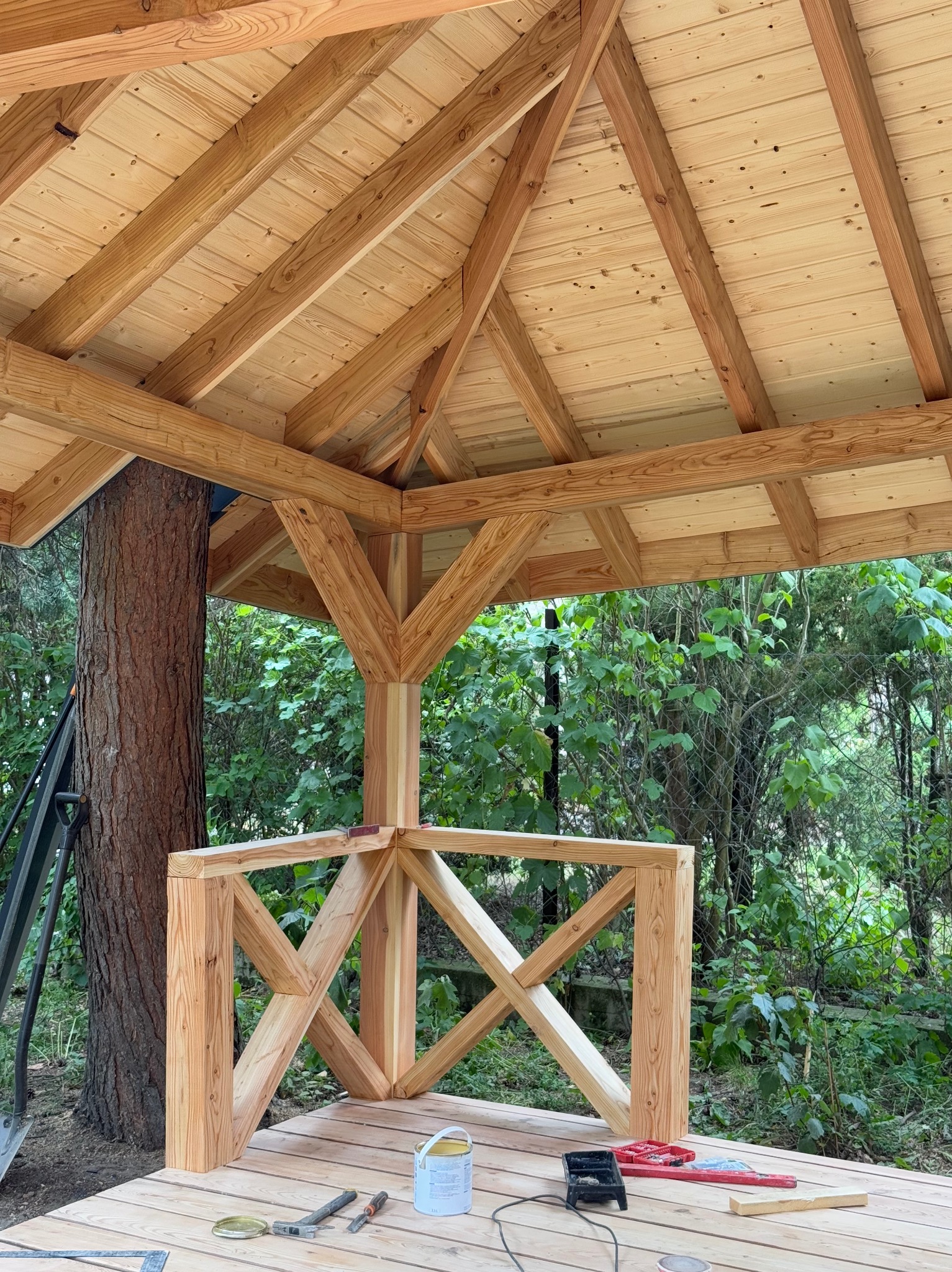 Drewniana altana w trakcie budowy, widoczne elementy konstrukcyjne dachu i balustrady, narzędzia na podłodze, drzewo wkomponowane w konstrukcję.